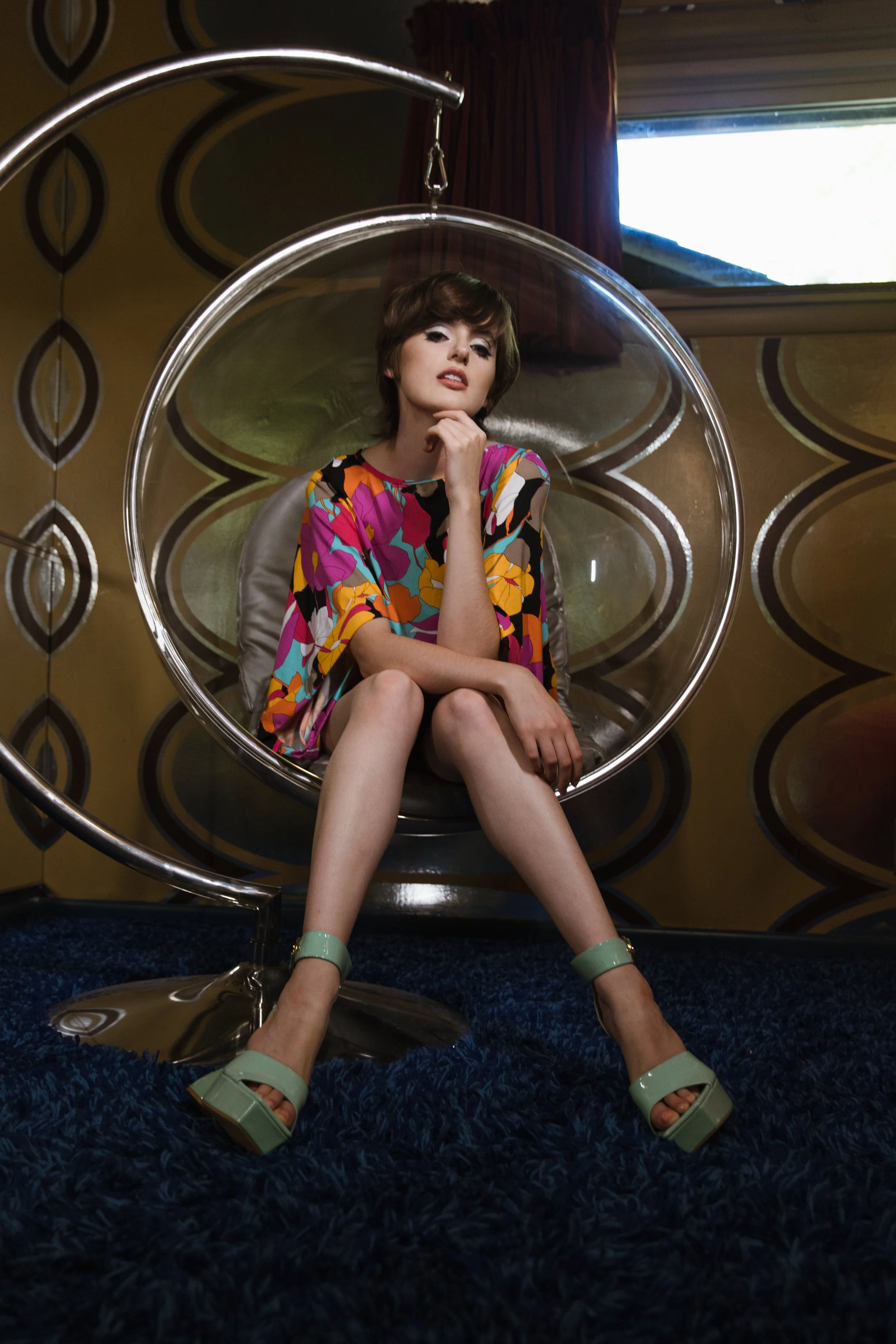 A woman with short brown hair and light skin sitting in a modern hanging chair with clear plastic and metal frame, dressed in a colorful patterned dress and green platform heels, in a room with retro-style patterned wallpaper and a window with red cu