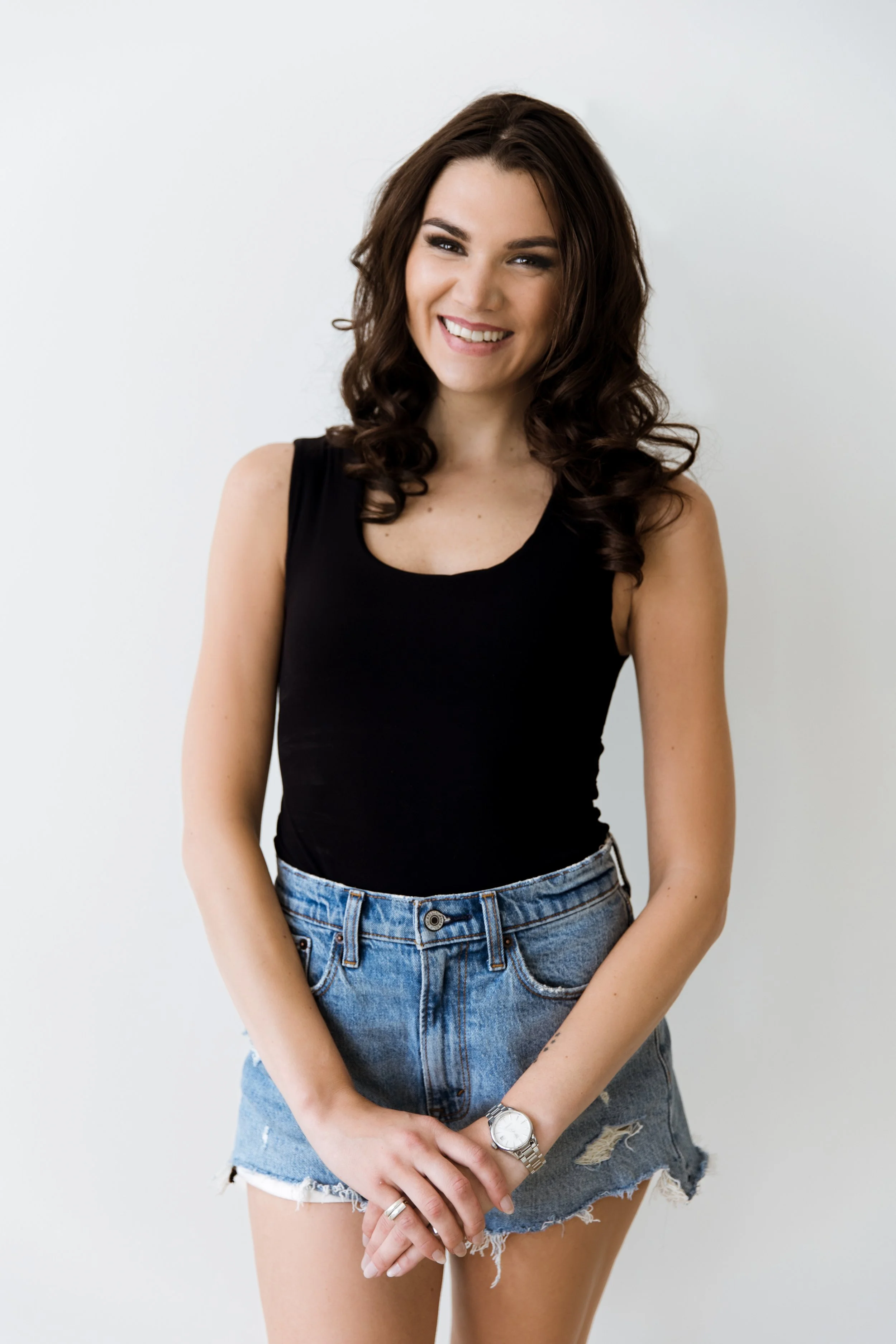 A young woman with brown curly hair, wearing a black sleeveless top and ripped denim shorts, smiling and standing against a plain white background.