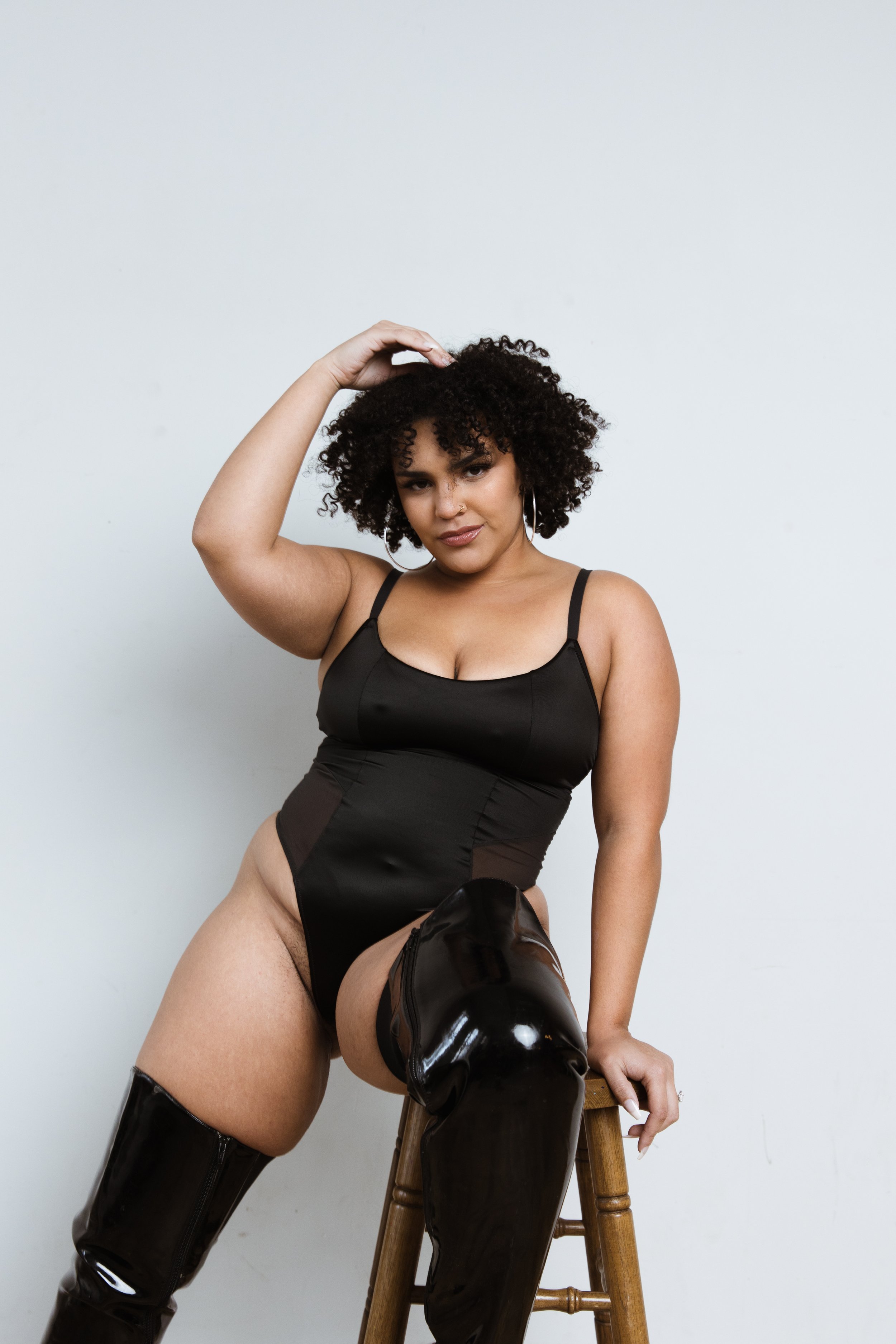 Woman with curly hair wearing black outfit and shiny black thigh-high boots posing against a plain white background.