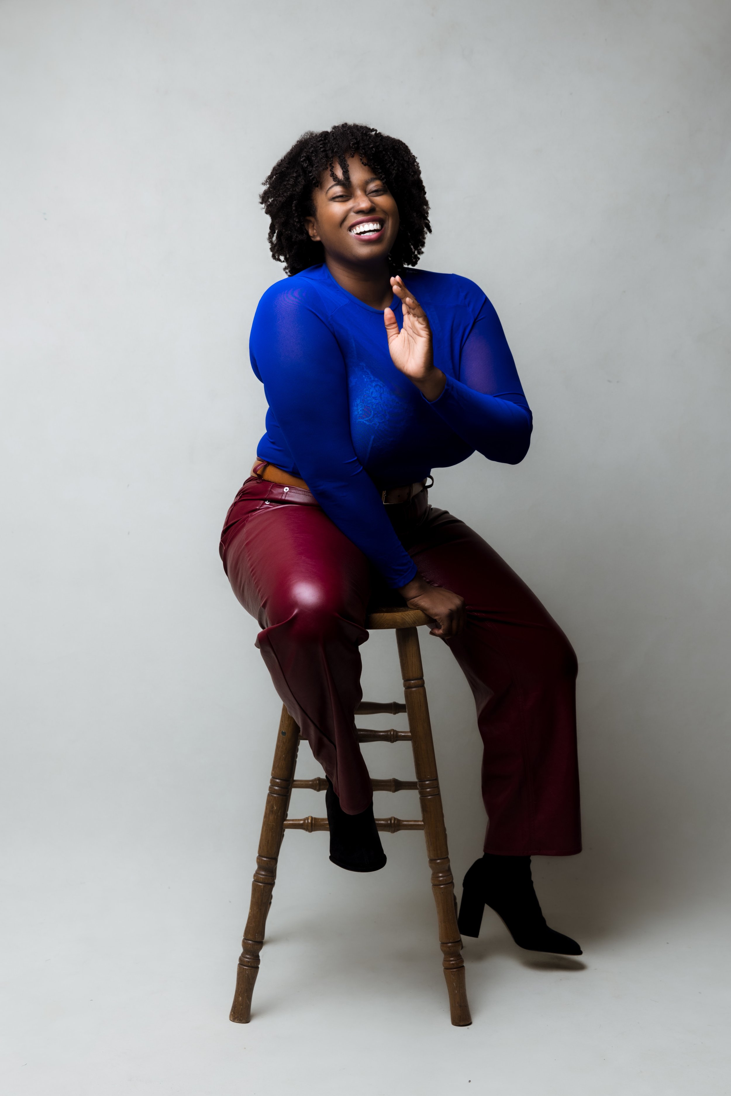 A woman sitting on a wooden stool, smiling and waving, wearing a blue long-sleeve top, maroon leather pants, and black high-heeled boots, against a plain light background.