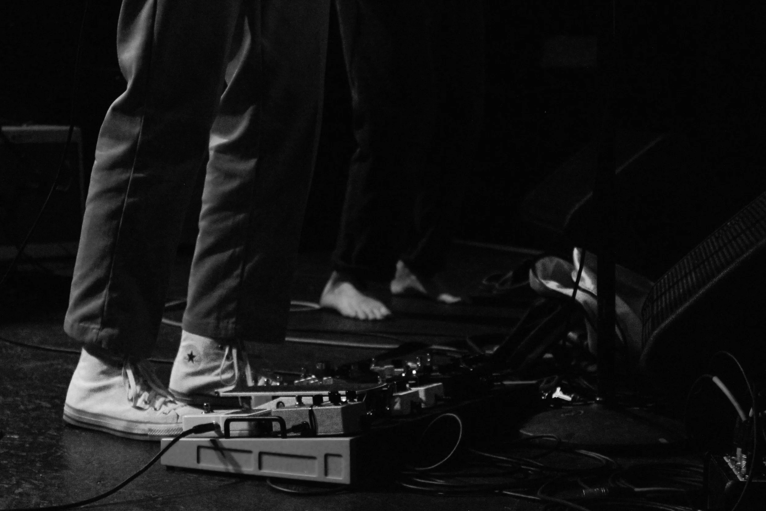Close-up of a person's legs and feet on a stage, with one foot on a pedalboard and the other foot barefoot, surrounded by audio equipment and cables, in black and white.