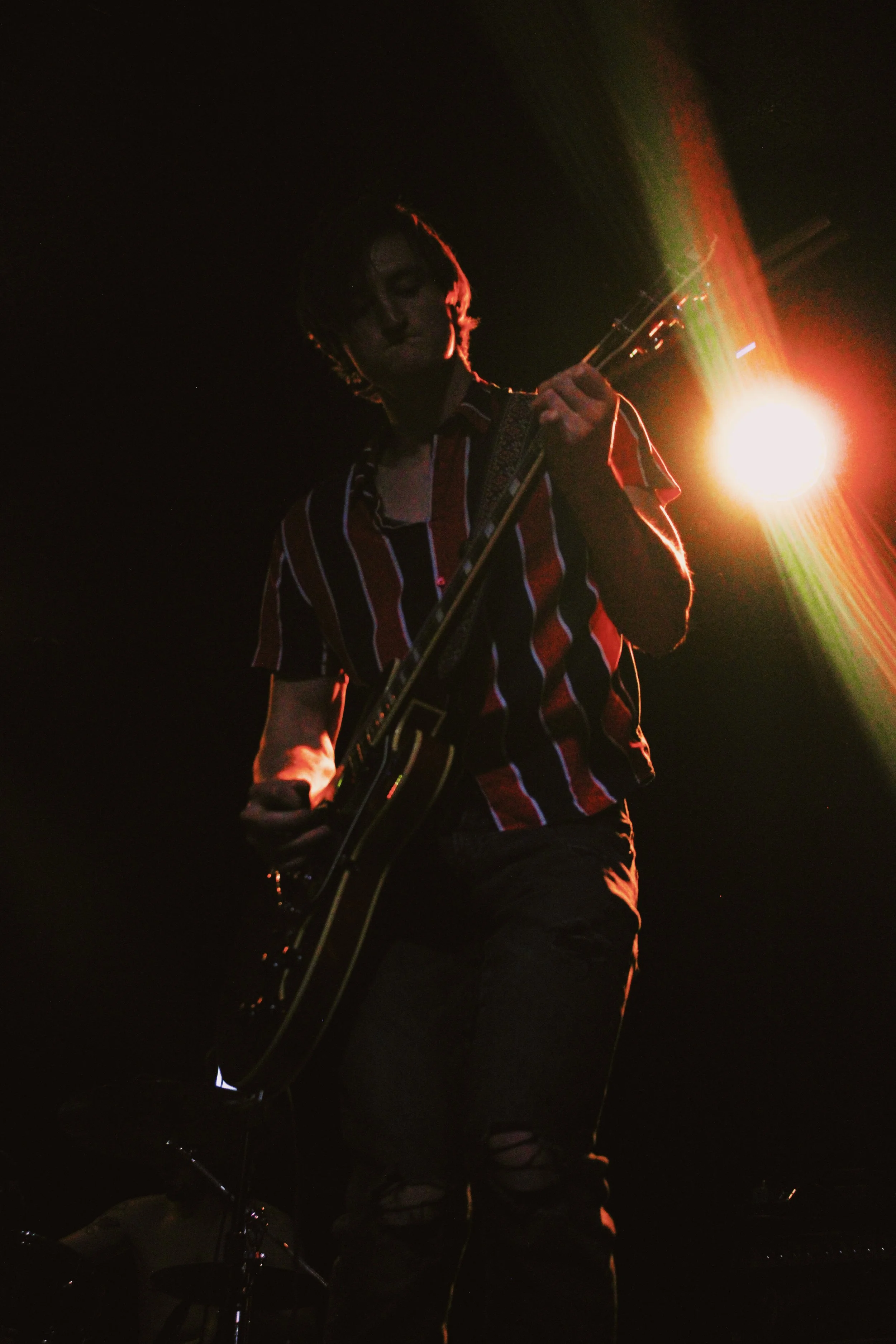 A person playing an electric guitar on stage, illuminated by bright stage lights, with a dark background.