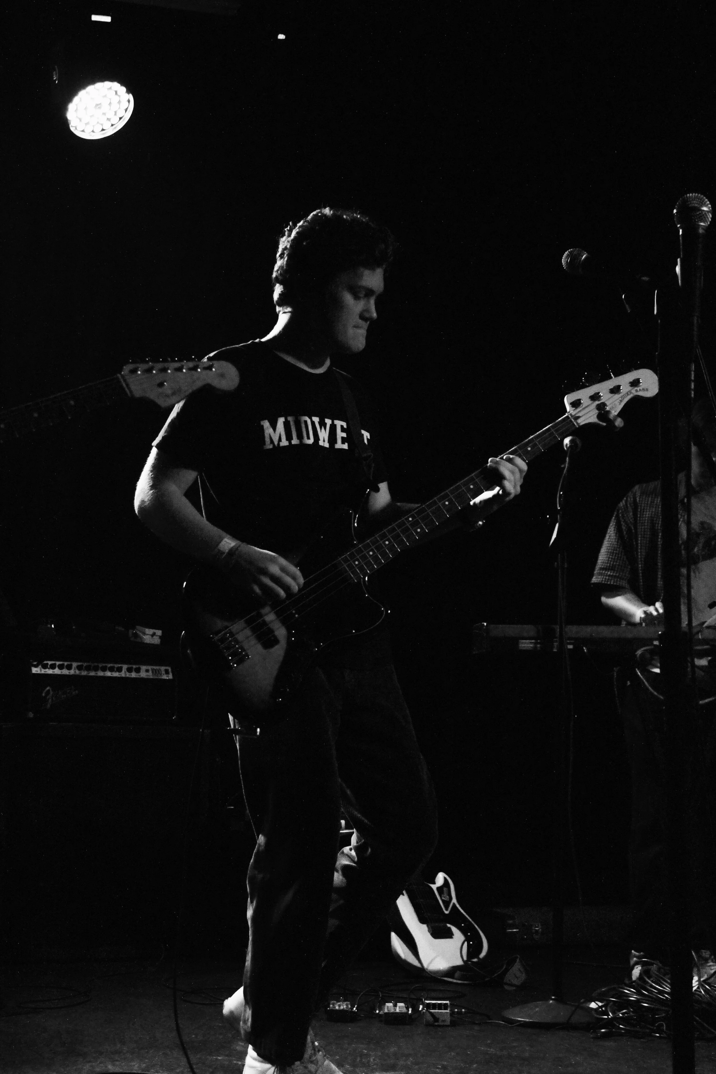 A young man playing an electric bass guitar on stage during a live music performance, in black and white.