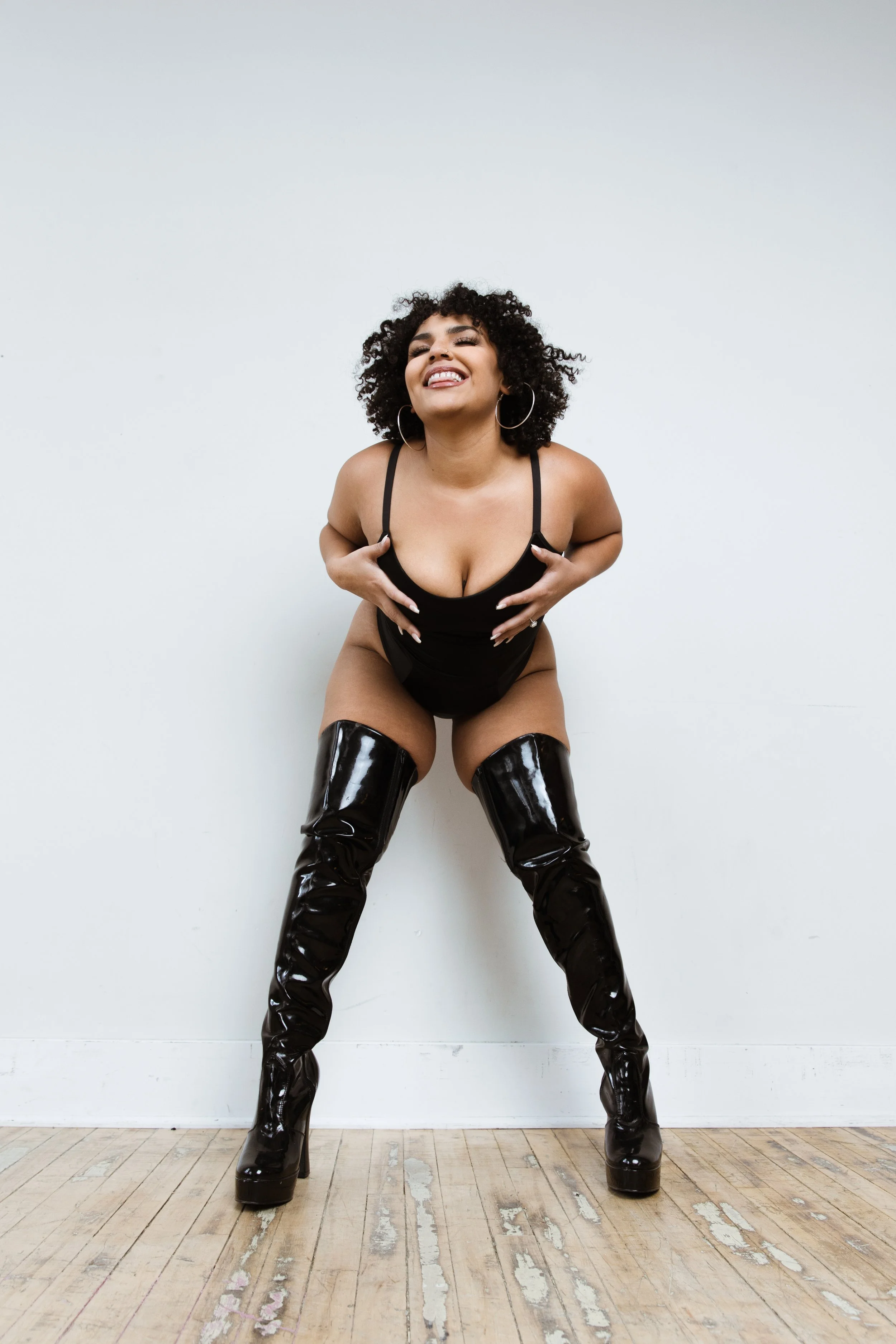 Woman with curly hair smiling, wearing black lingerie and thigh-high black shiny boots, standing against a white wall in a playful pose.