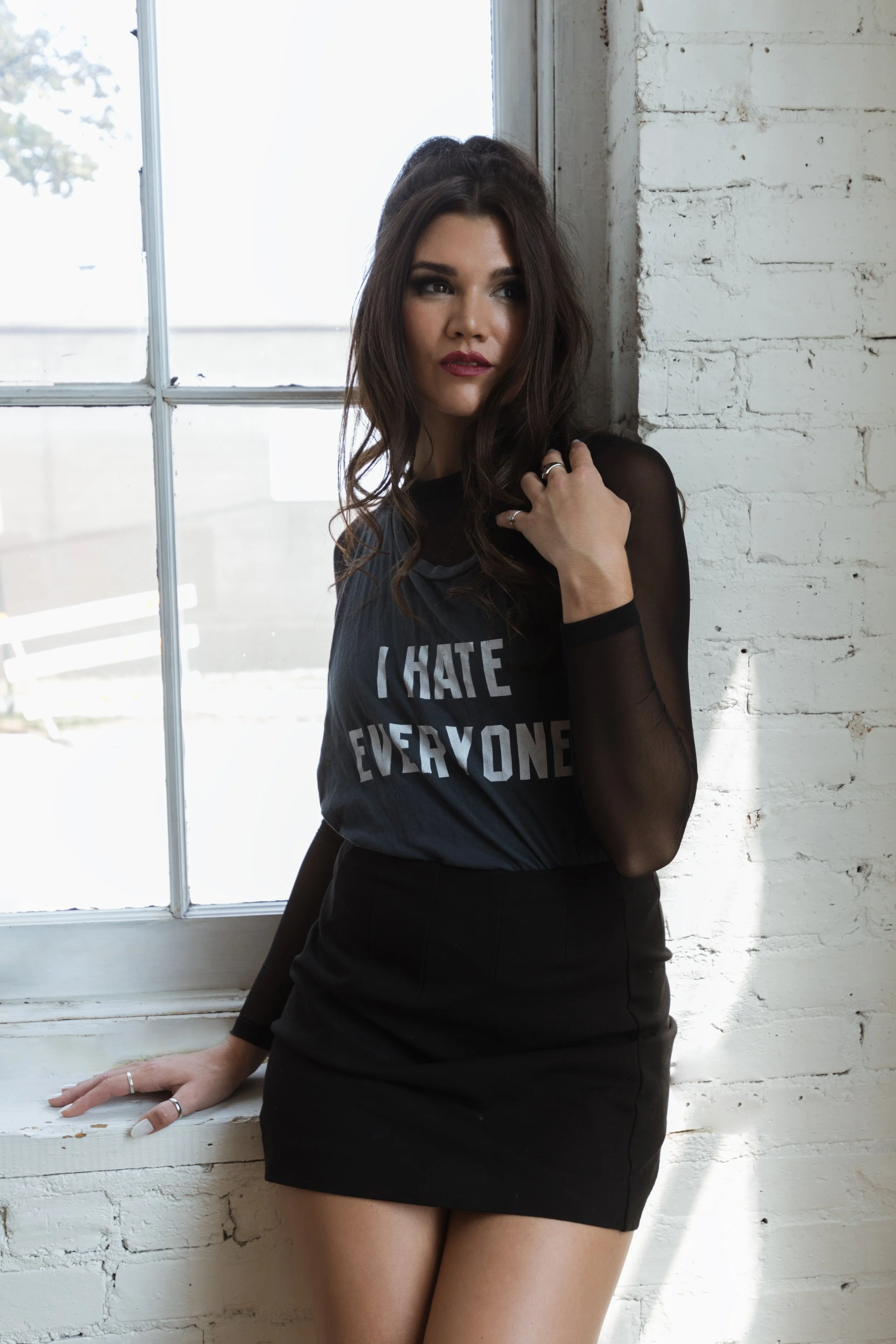 A woman with long dark hair stands by a white brick wall next to a window, wearing a black t-shirt with the text 'I HATE EVERYONE' and a black skirt, with natural light illuminating her face.
