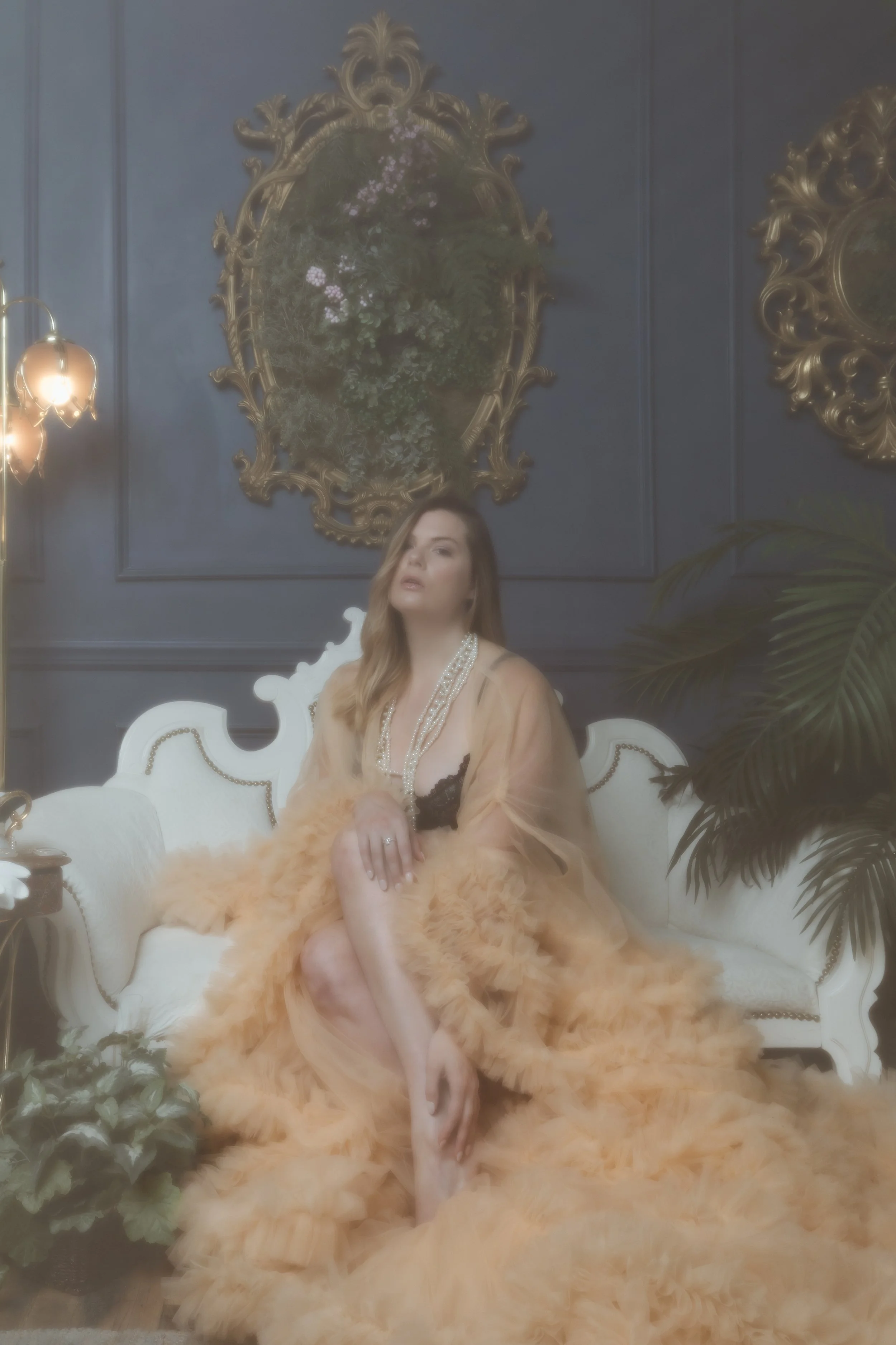 A woman in a peach-colored, feathered gown with black lace accents, wearing pearls, seated on a vintage white sofa in an opulent room with dark blue walls, gold ornate mirrors, and lush green plants.