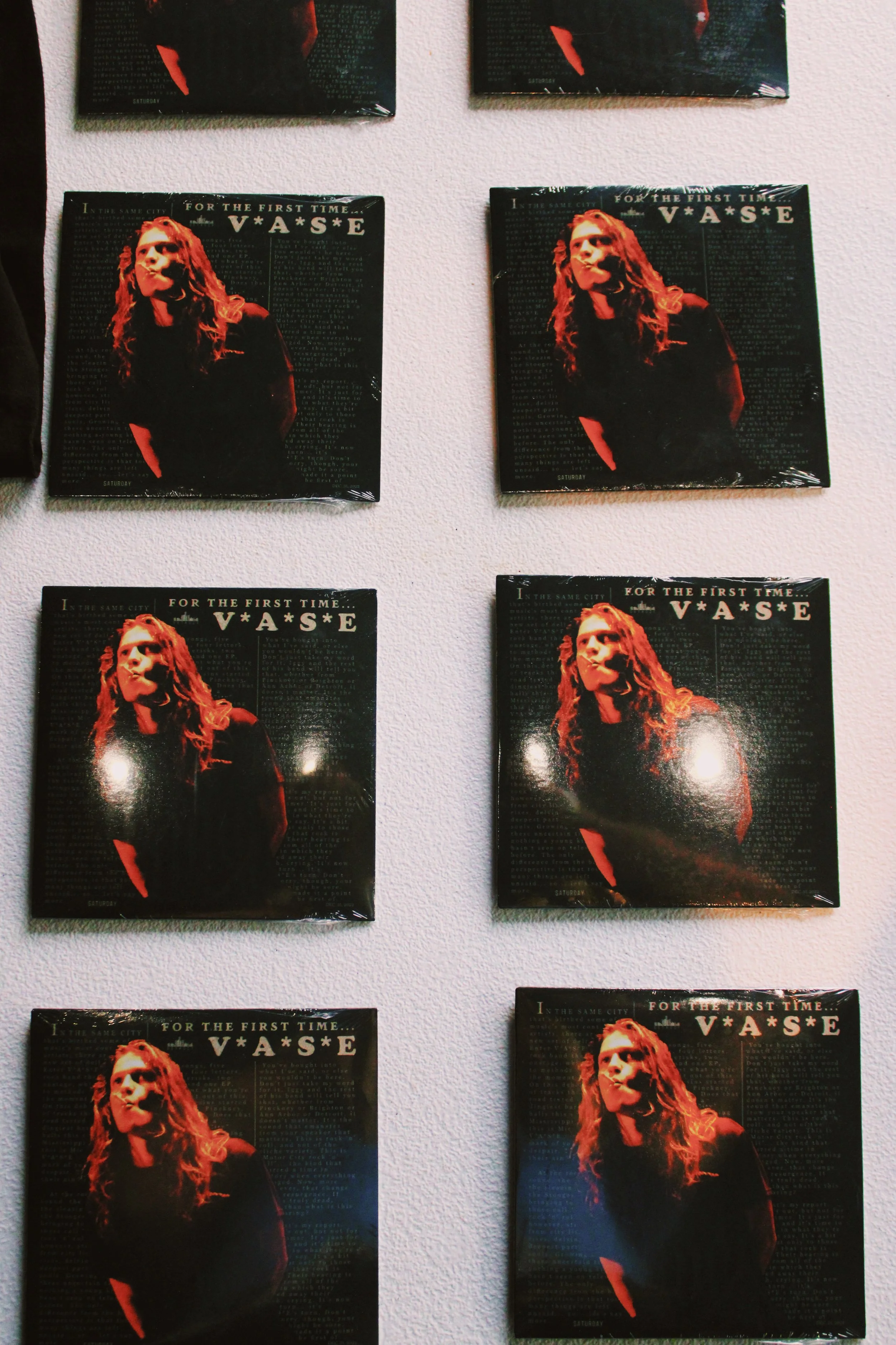Nine posters of a woman with long, curly hair, wearing dark clothing, and making a puffed-cheek expression, arranged on a white wall in a 3x3 grid. The posters feature text that says "For the first time... V*A*S*E" and additional smaller print.