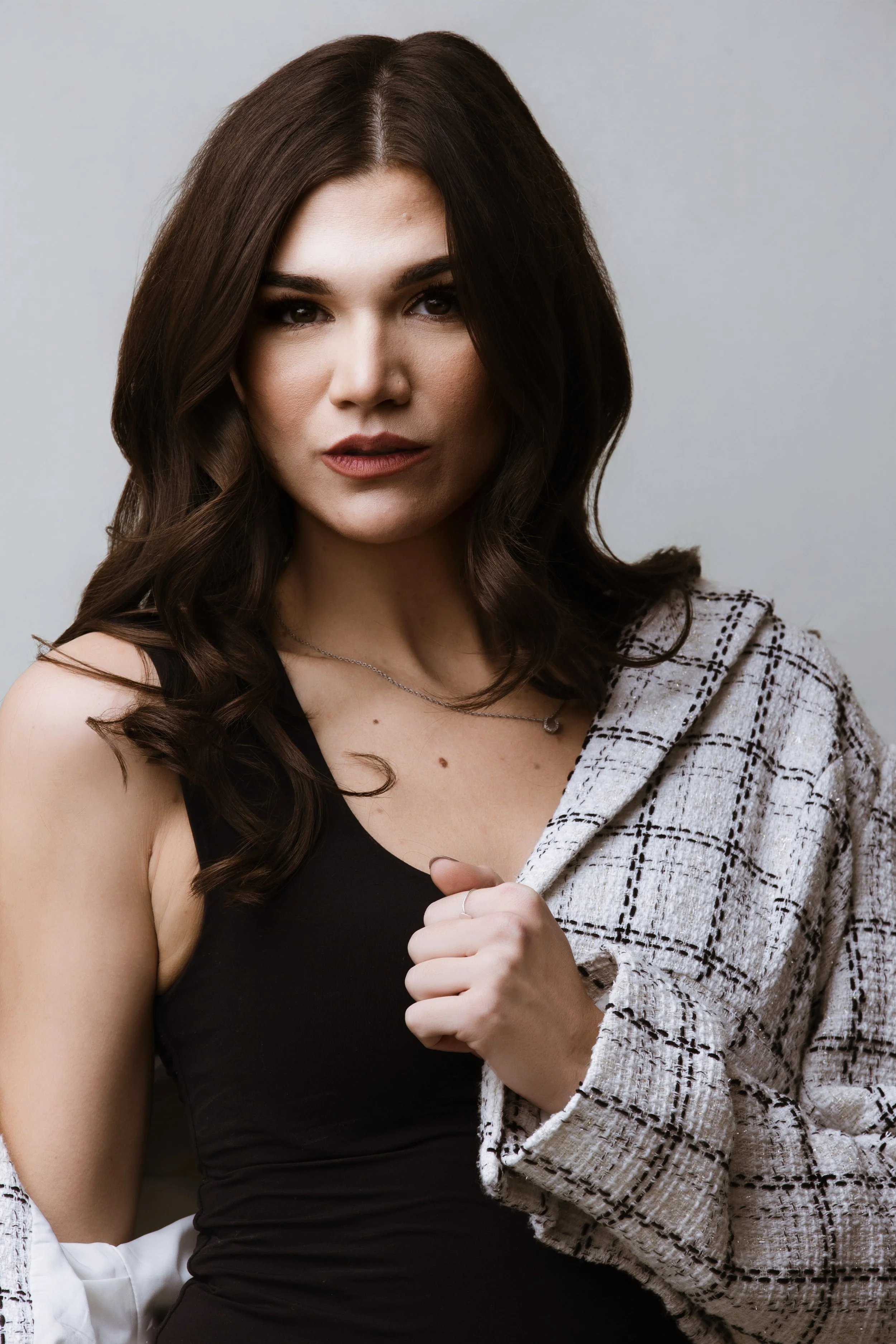 A woman with long brown hair, wearing a black sleeveless top and a light plaid jacket, looking confidently at the camera.