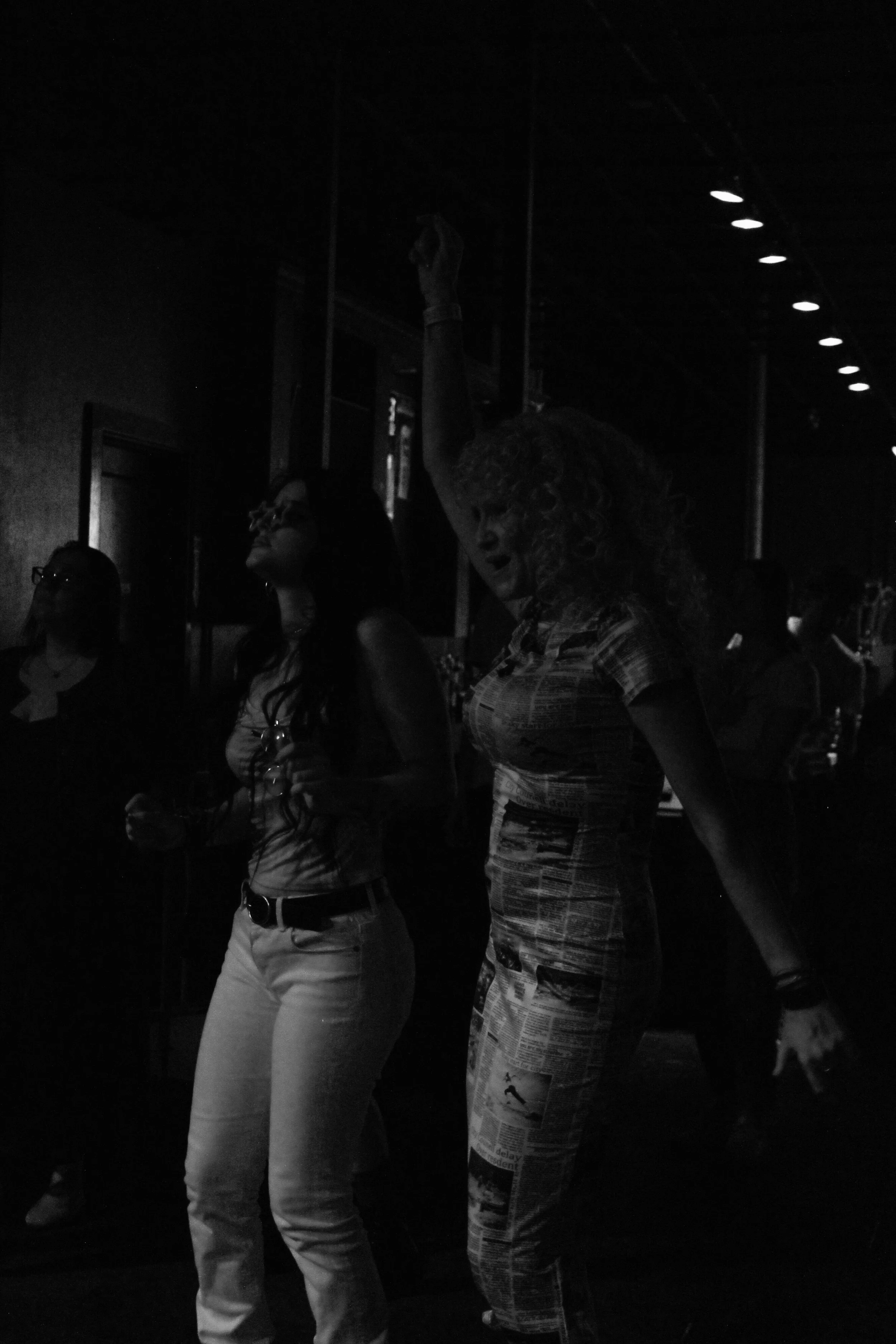 Two women at a bar or nightclub, one dancing with her arm raised and wearing a patterned outfit, the other holding a drink and wearing glasses and a white top, with other people in the background in a dark setting with ceiling lights.