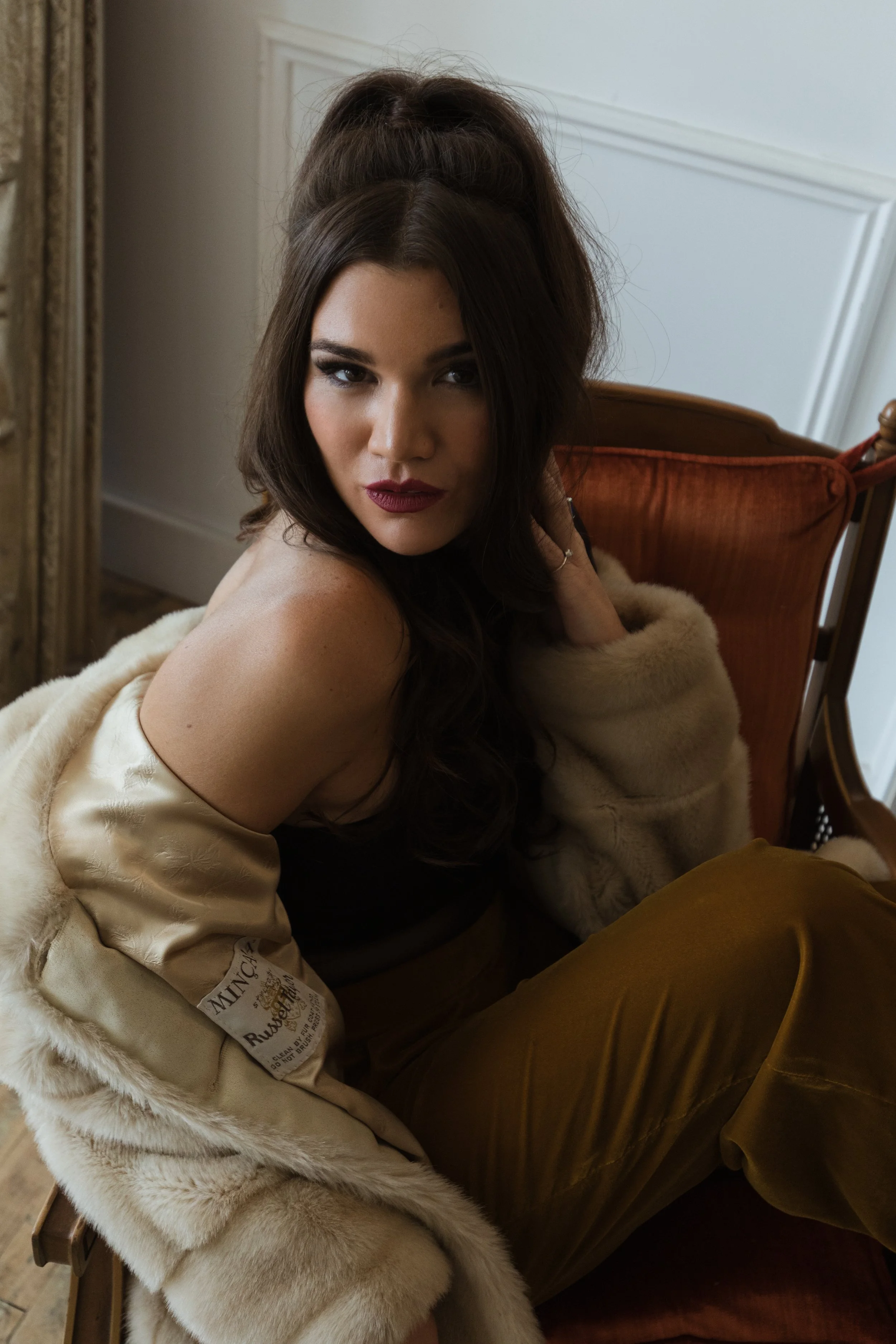 A woman with dark wavy hair and dark lipstick sitting on an orange armchair, wearing a beige fur coat and gold satin pants, looking at the camera.