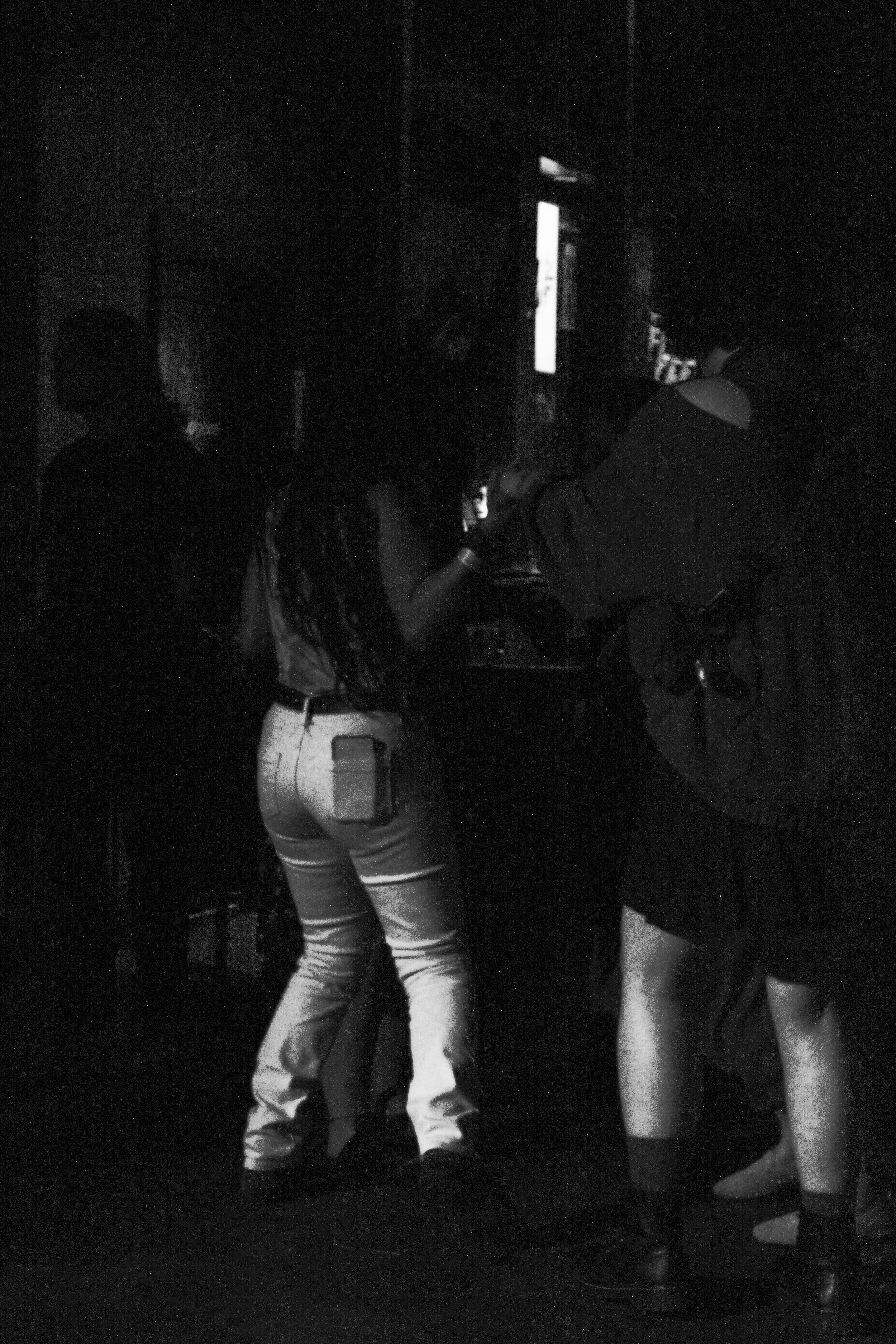 People standing and talking in a dark indoor setting, illuminated by a screen or monitor.