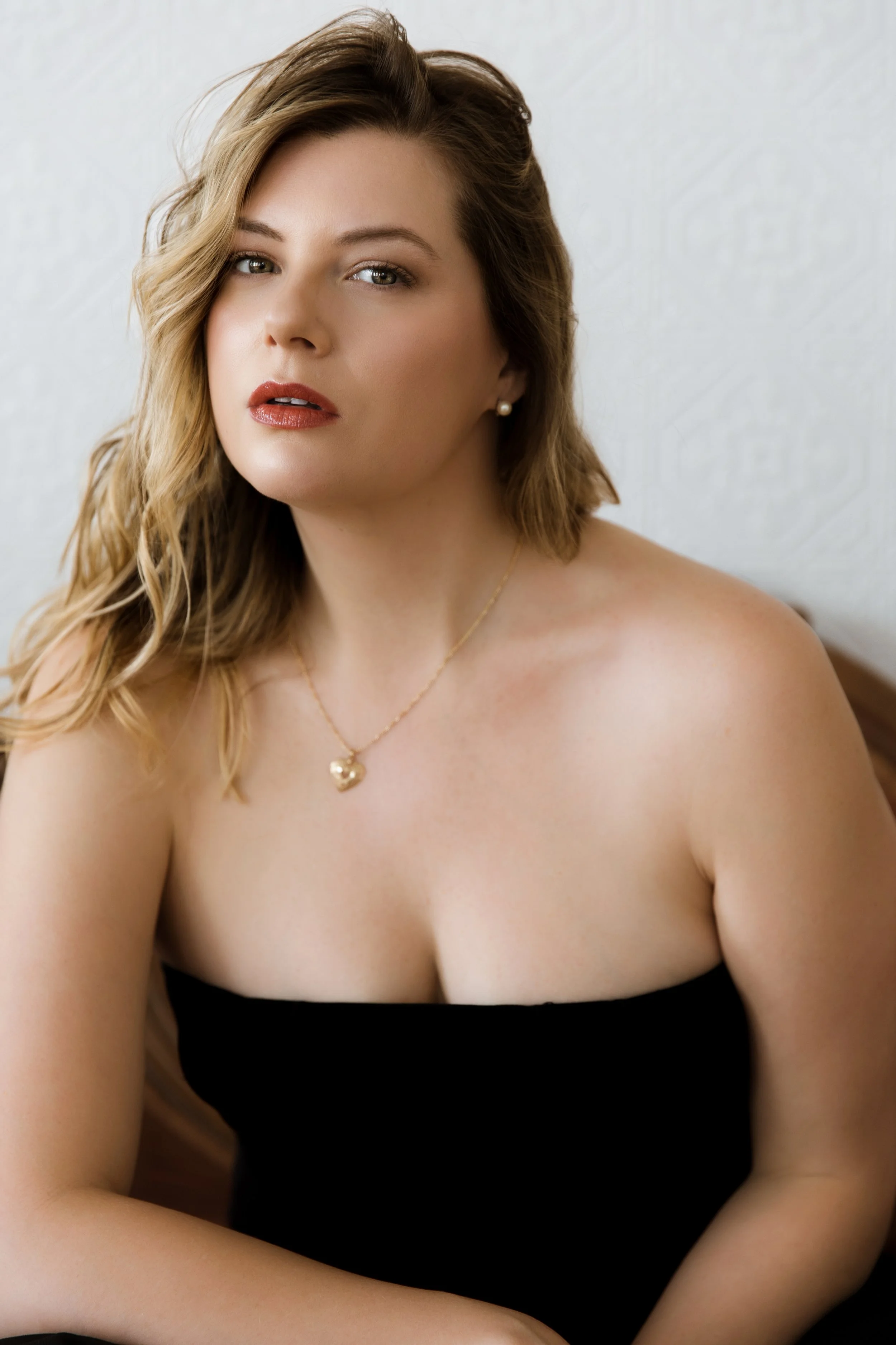 A woman with wavy, shoulder-length hair, wearing a black strapless top, gold necklace with a heart-shaped pendant, and pearl earrings, sitting against a light-colored wall.
