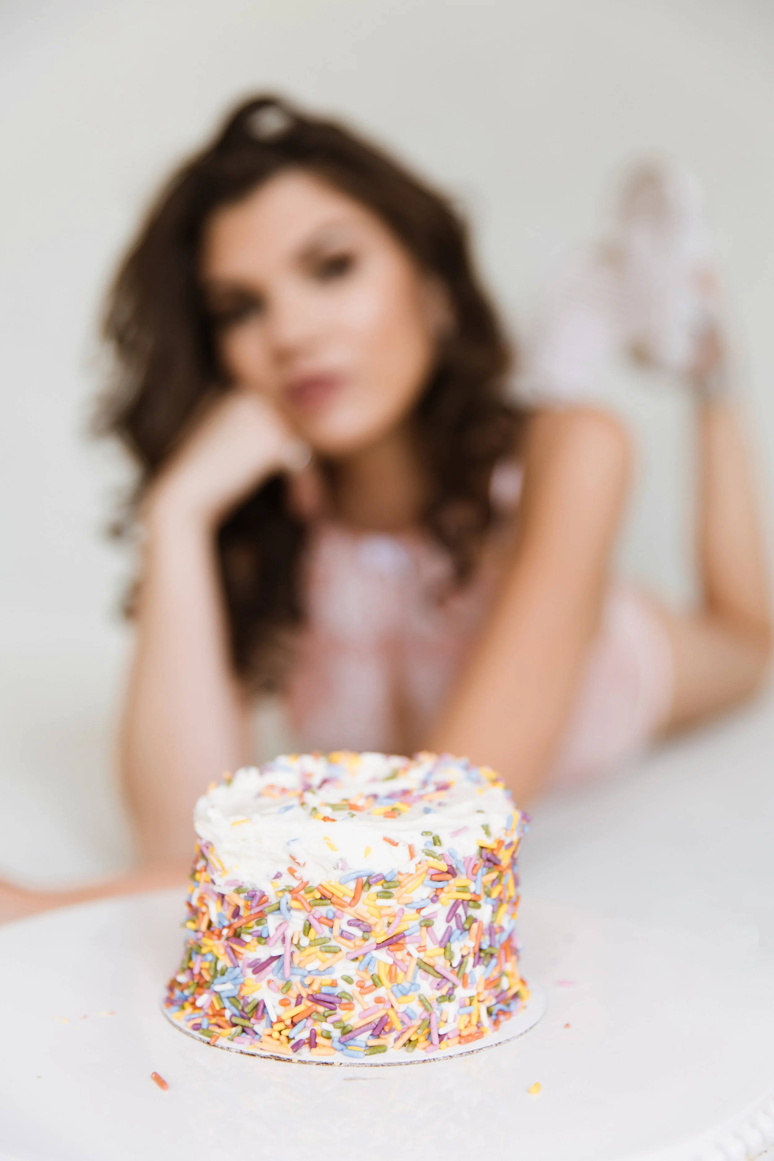 A woman lying on a white surface with a colorful sprinkle-covered birthday cake in the foreground.