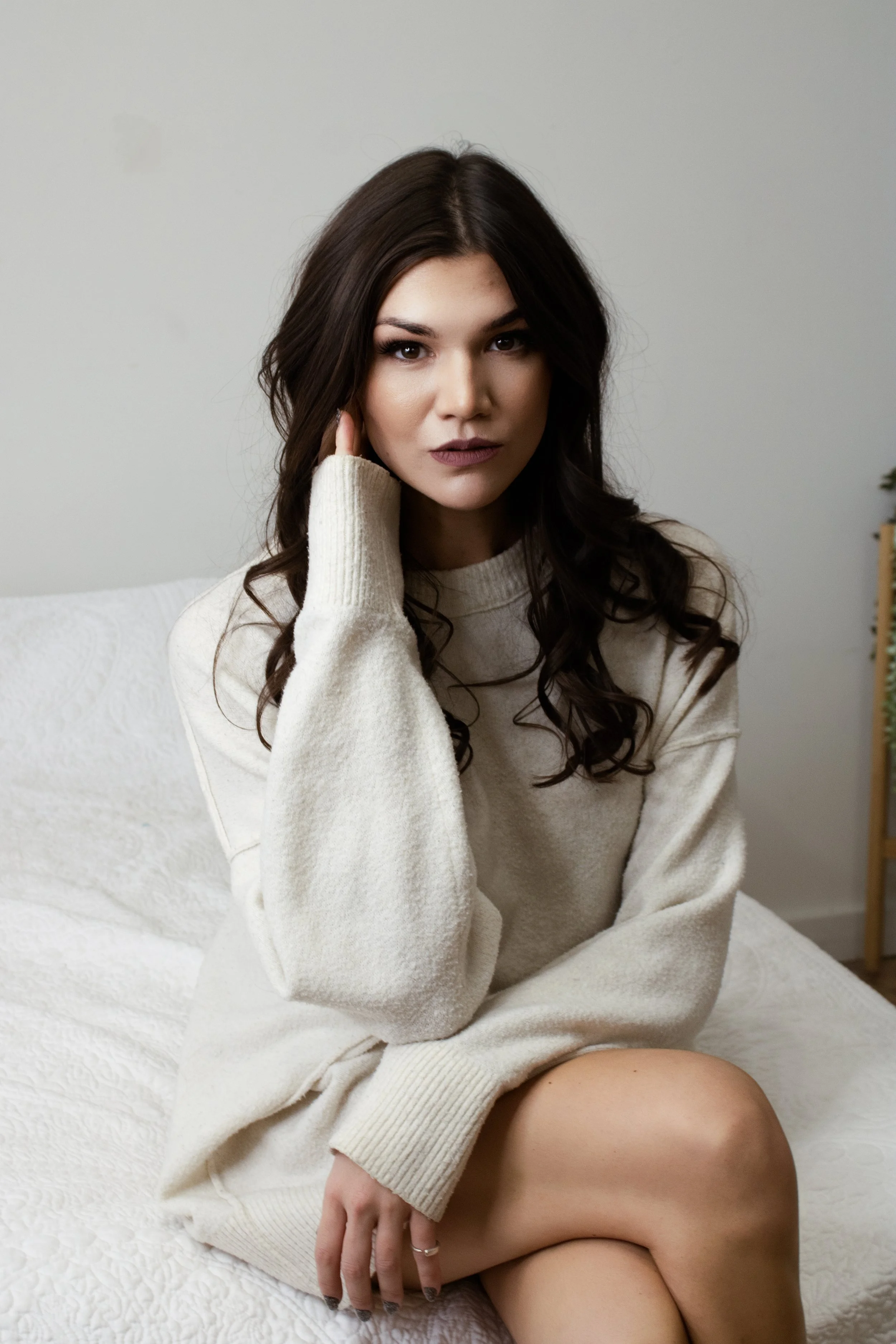 A young woman with dark curly hair sitting on a bed, wearing a cream-colored sweater, looking directly at the camera with a neutral expression.