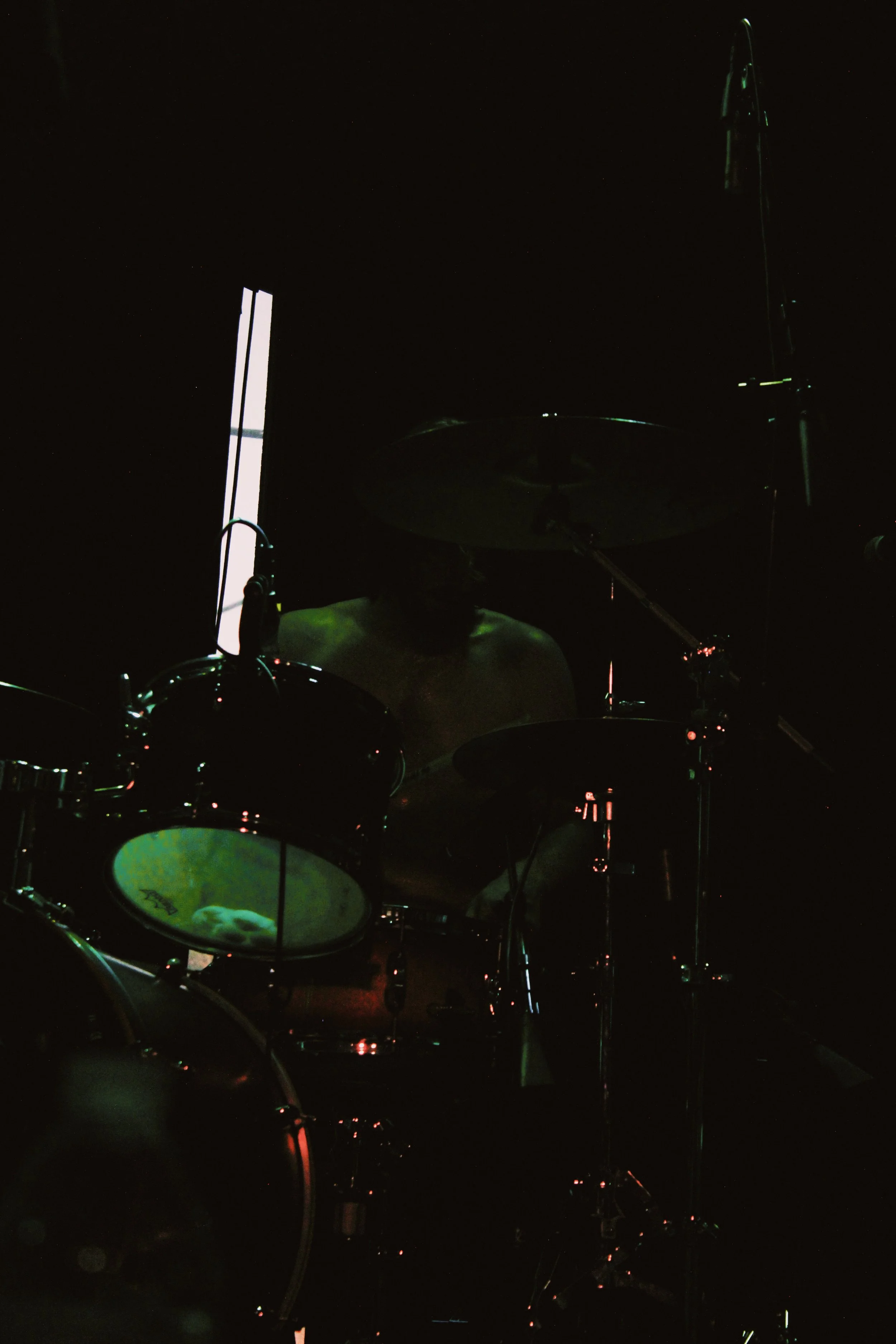 Dark image of a shirtless man playing a drum set, with a bright vertical light in the background.