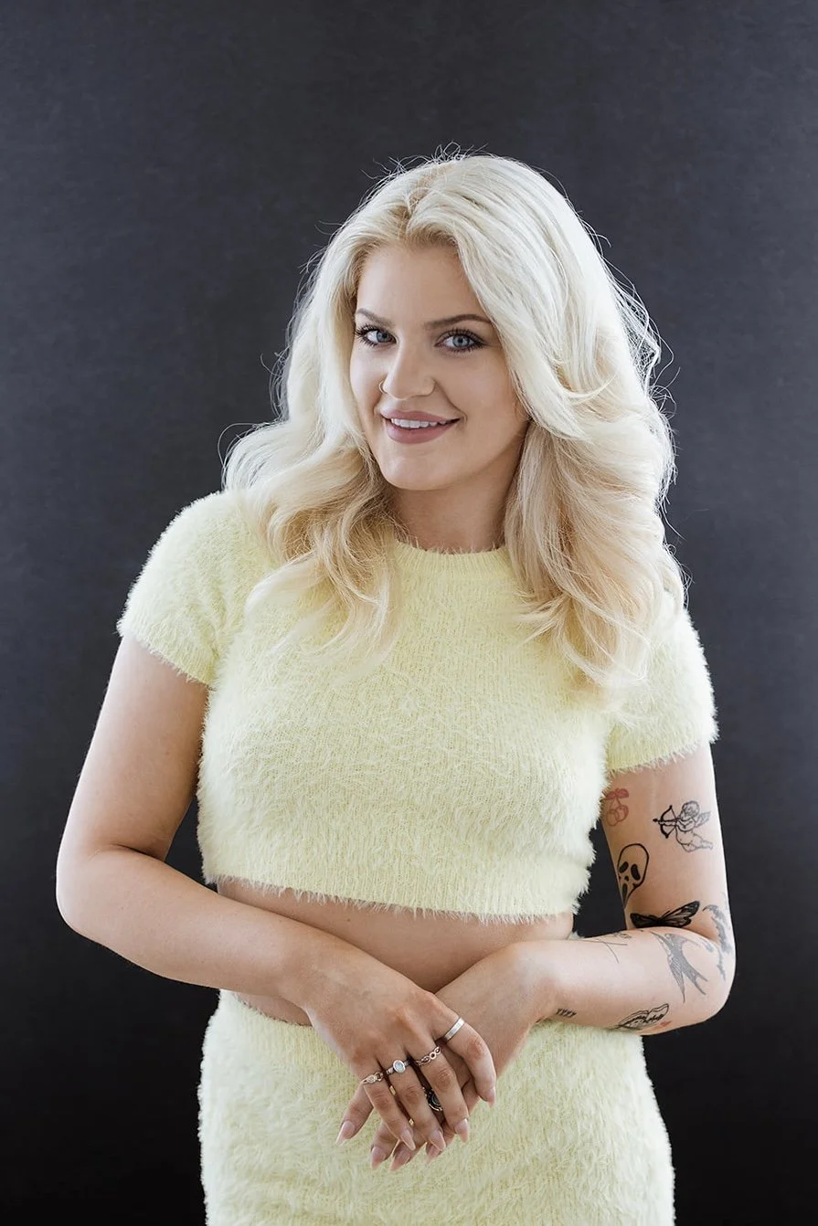 A young woman with platinum blonde hair styled in loose waves, wearing a pale yellow fuzzy crop top and matching skirt, smiling with her hands clasped in front of her, standing against a dark background.
