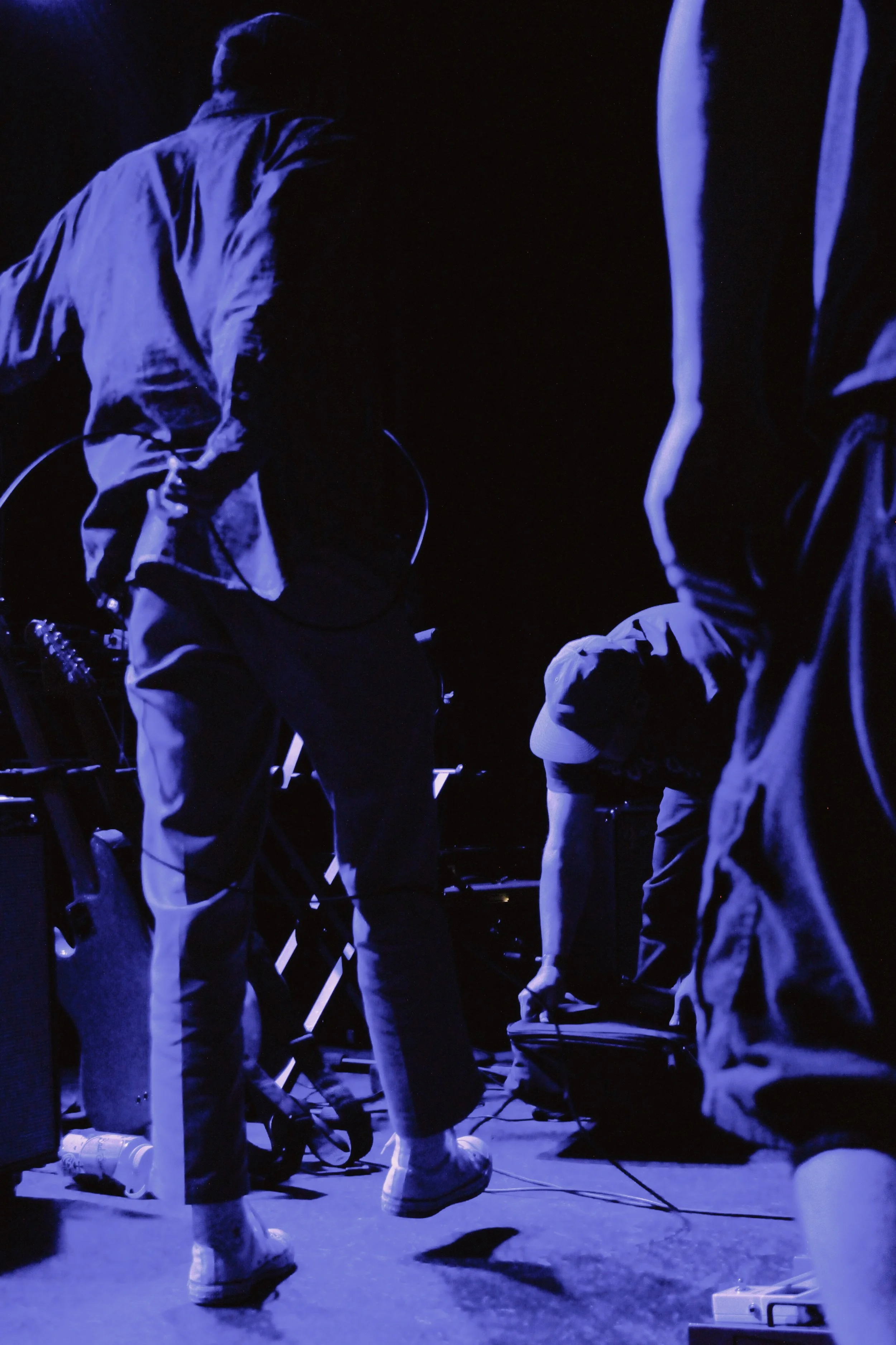 Two people on stage, one standing and the other bending down, under dramatic blue lighting, with musical equipment nearby.