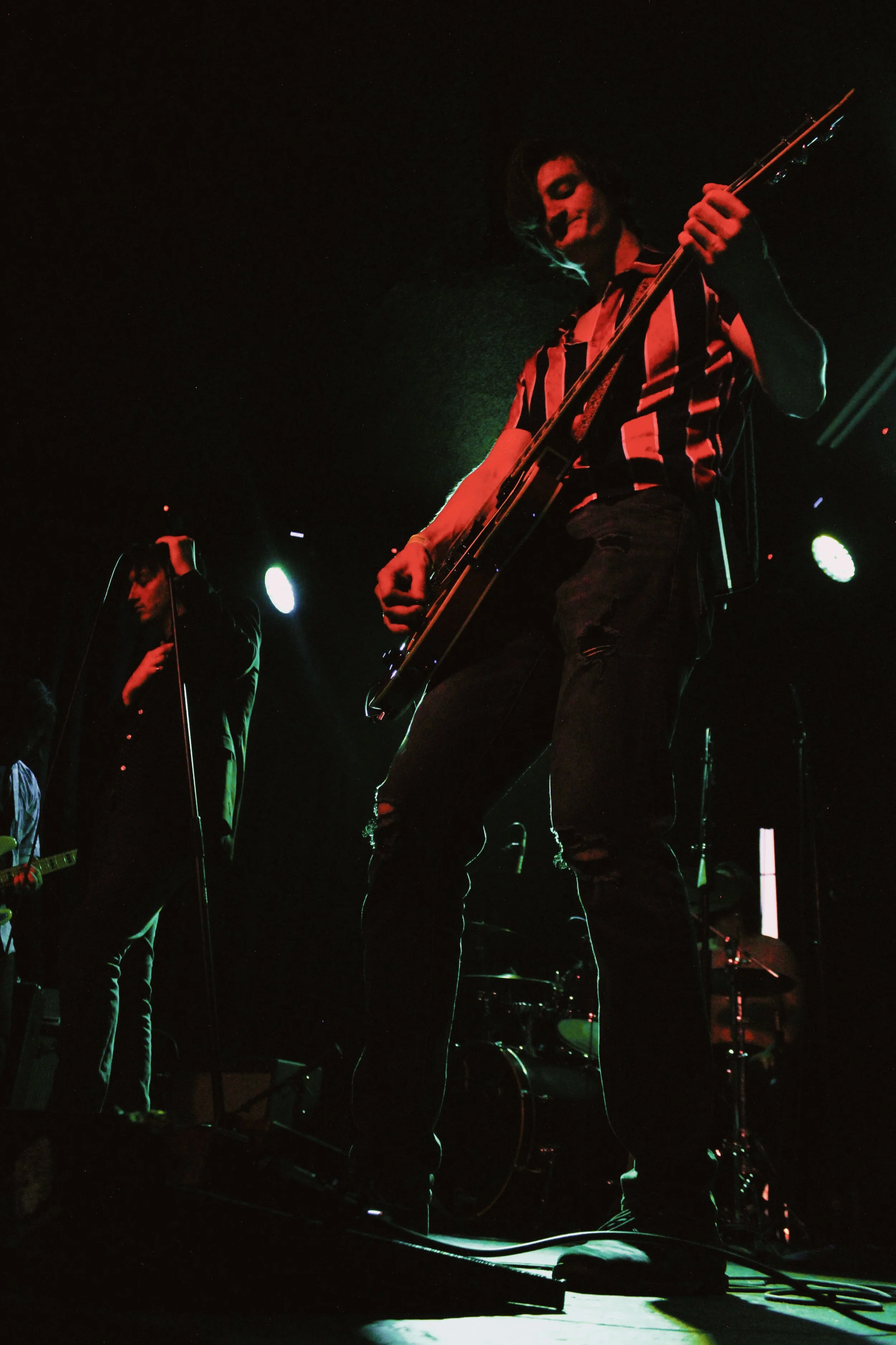 A musical band performing live on stage with a guitarist in the foreground playing with focus, in a dark setting with stage lights.
