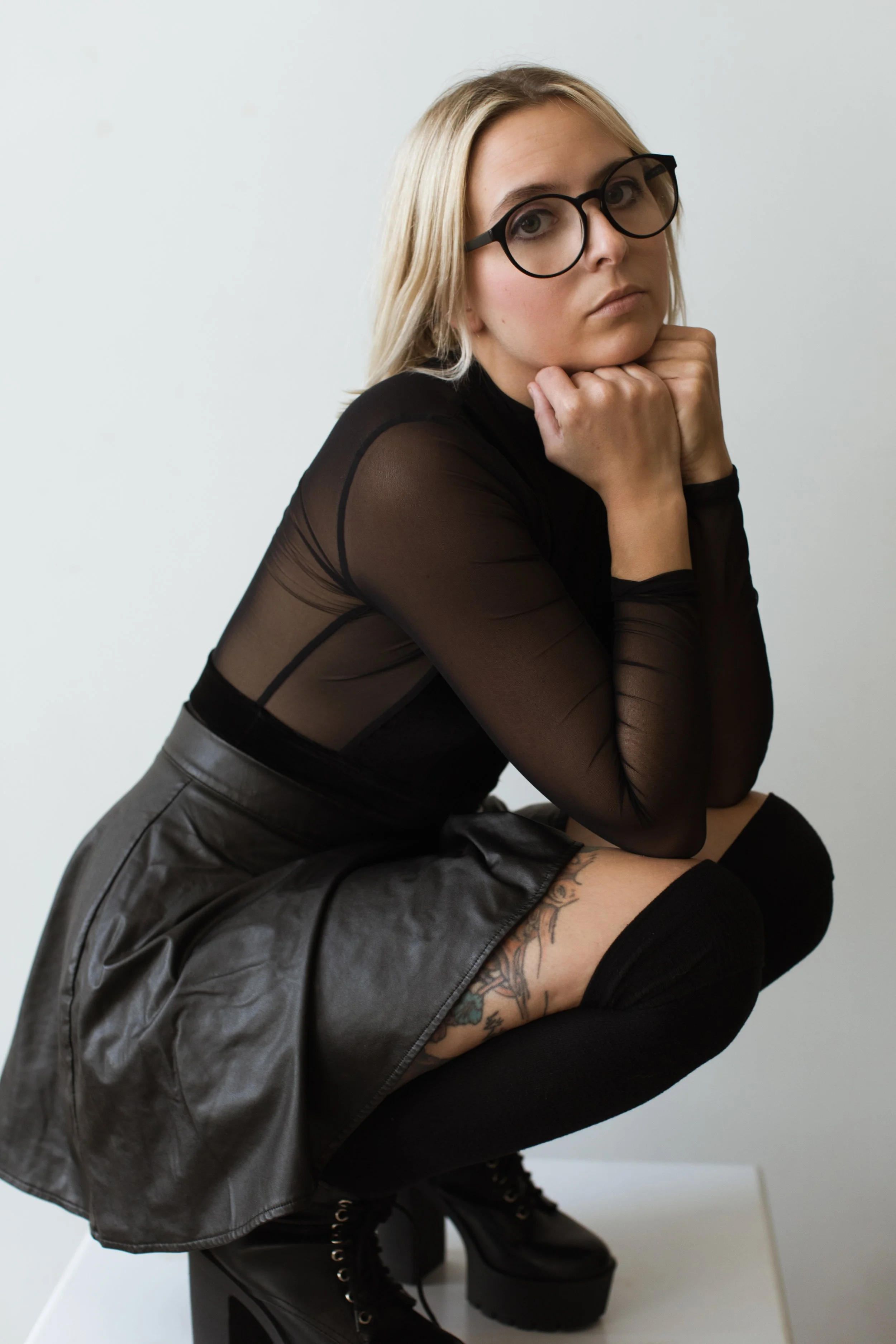 A young woman with blonde hair, glasses, and tattoos, wearing a black sheer top, leather skirt, and black boots, squatting with her chin resting on her hands against a plain white background.