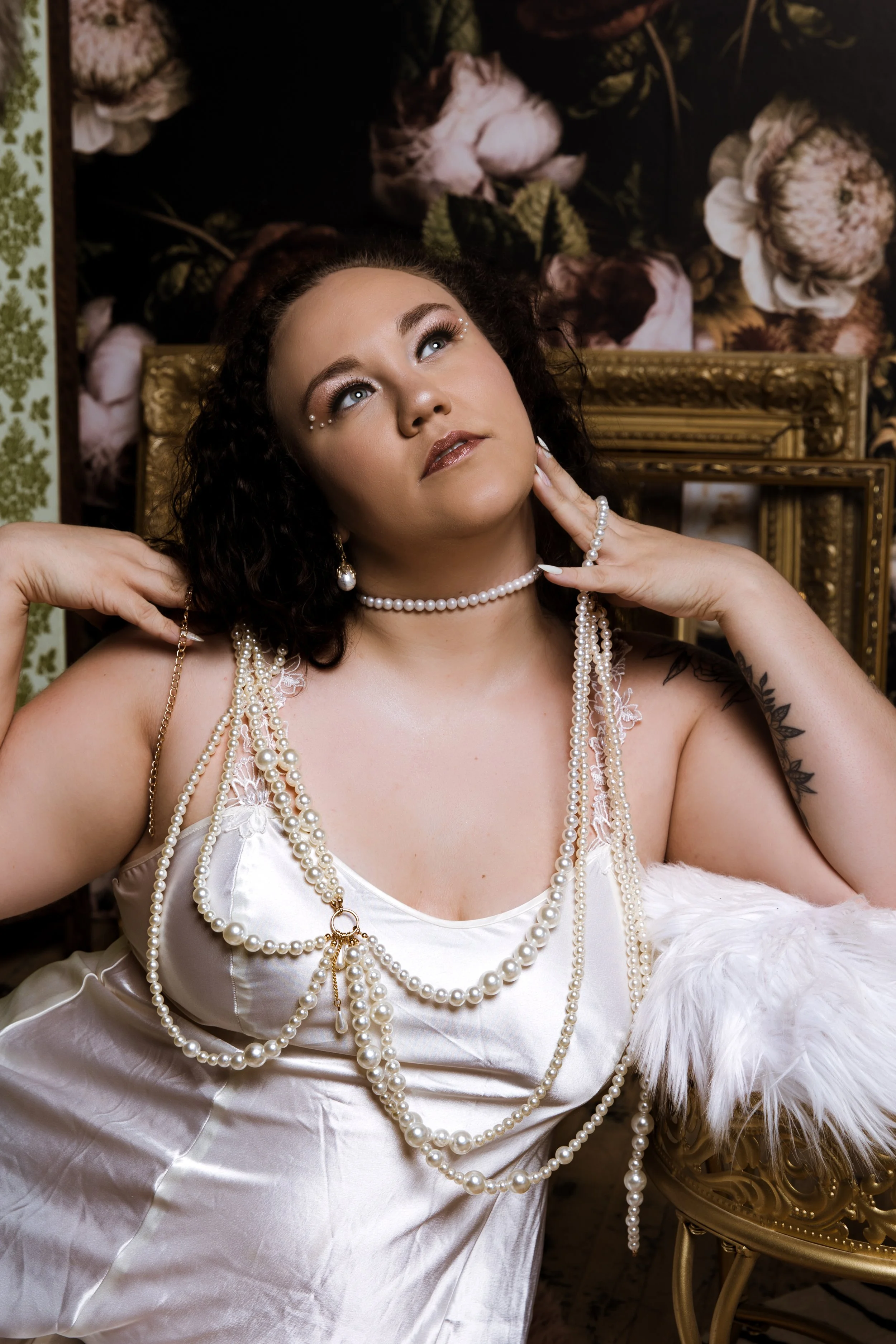 Woman with dark curly hair wearing multiple strands of pearl necklaces, pearl earrings, and a pearl choker, resting her head on her hand, seated in an ornate room with floral wallpaper and gold picture frames.