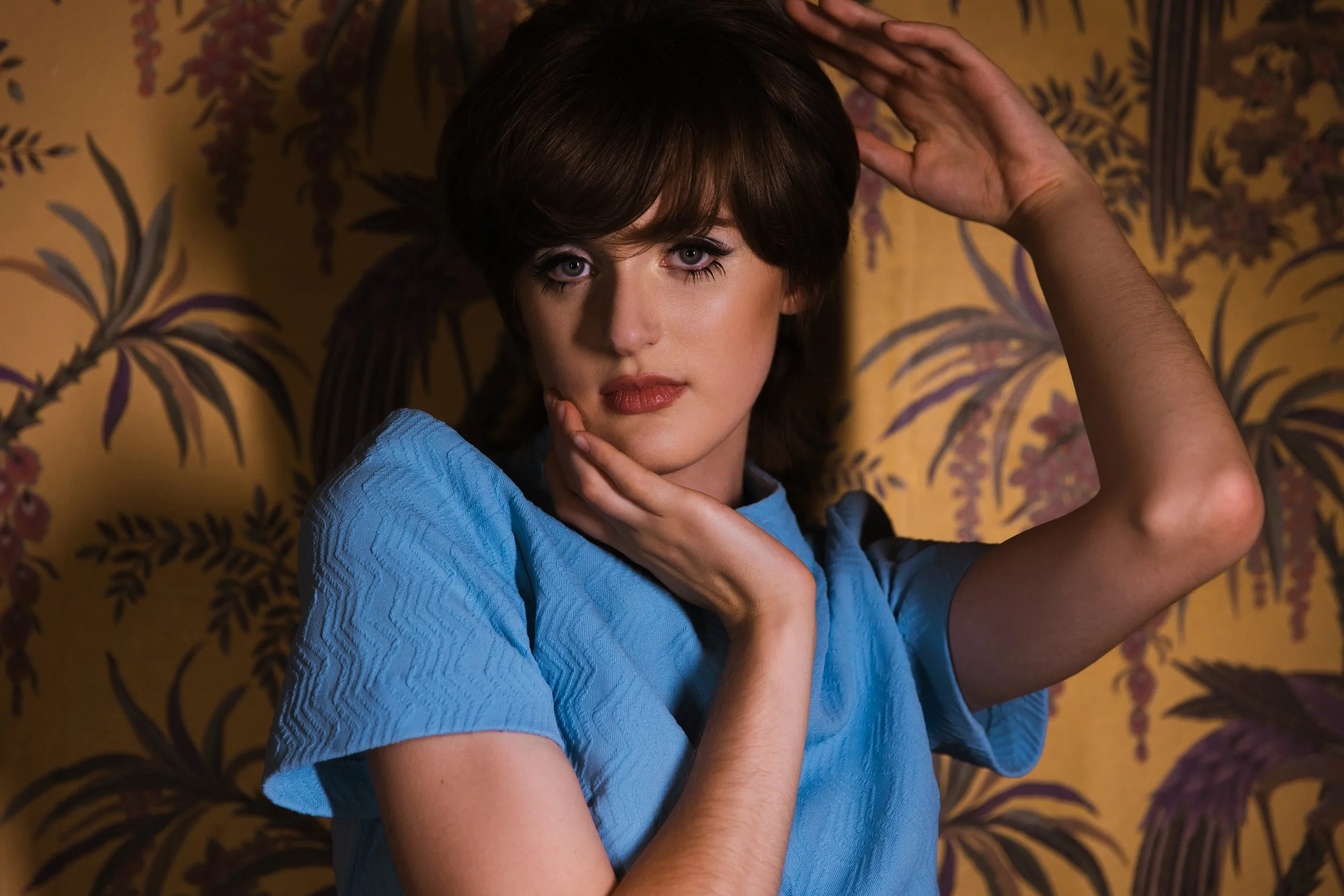A woman with short brown hair wearing a blue top, posing with one hand on her chin and the other on her head, against a yellow background with a floral pattern.