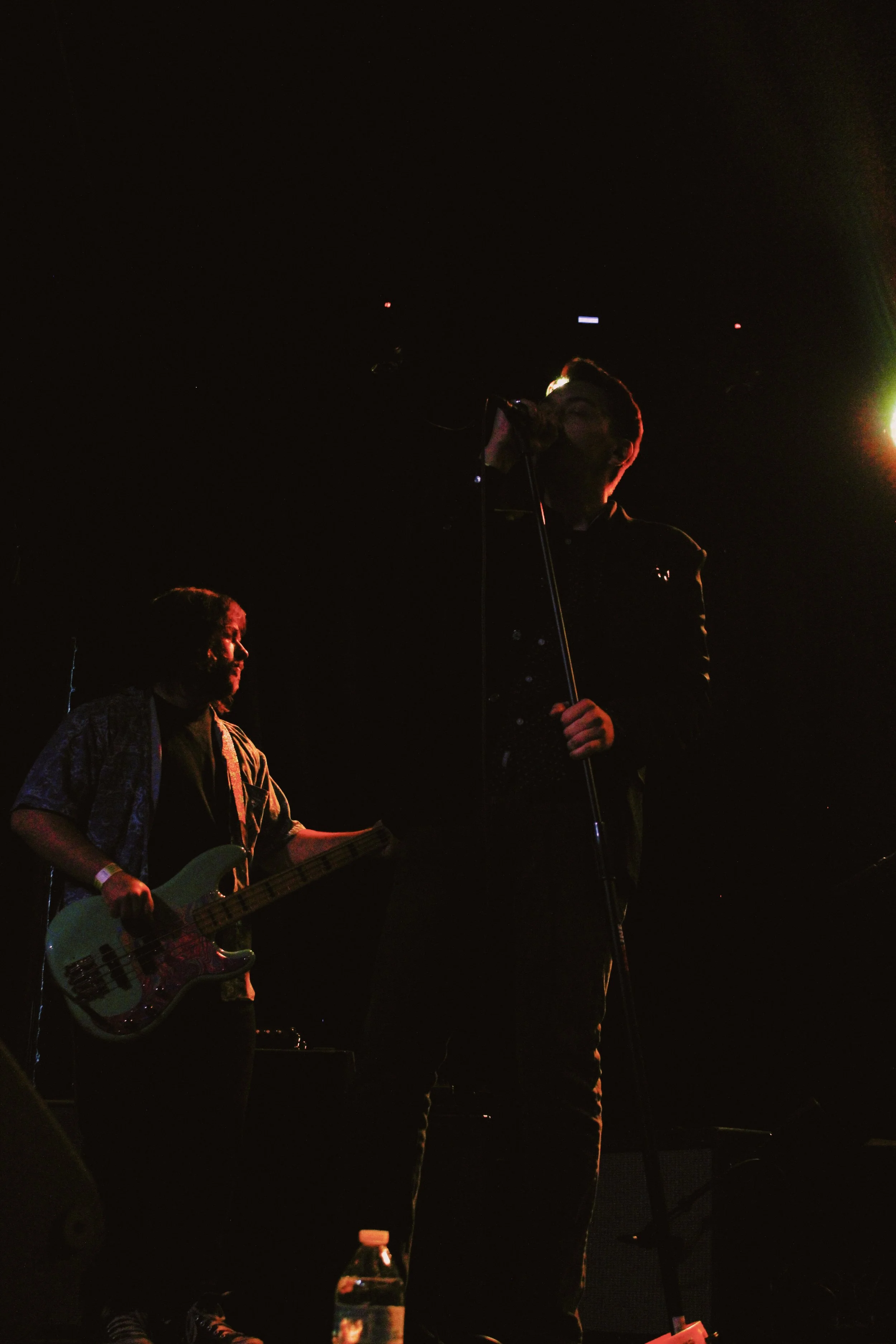 Two musicians performing on stage in darkness, one with a guitar and the other singing into a microphone, with a water bottle on the stage floor.