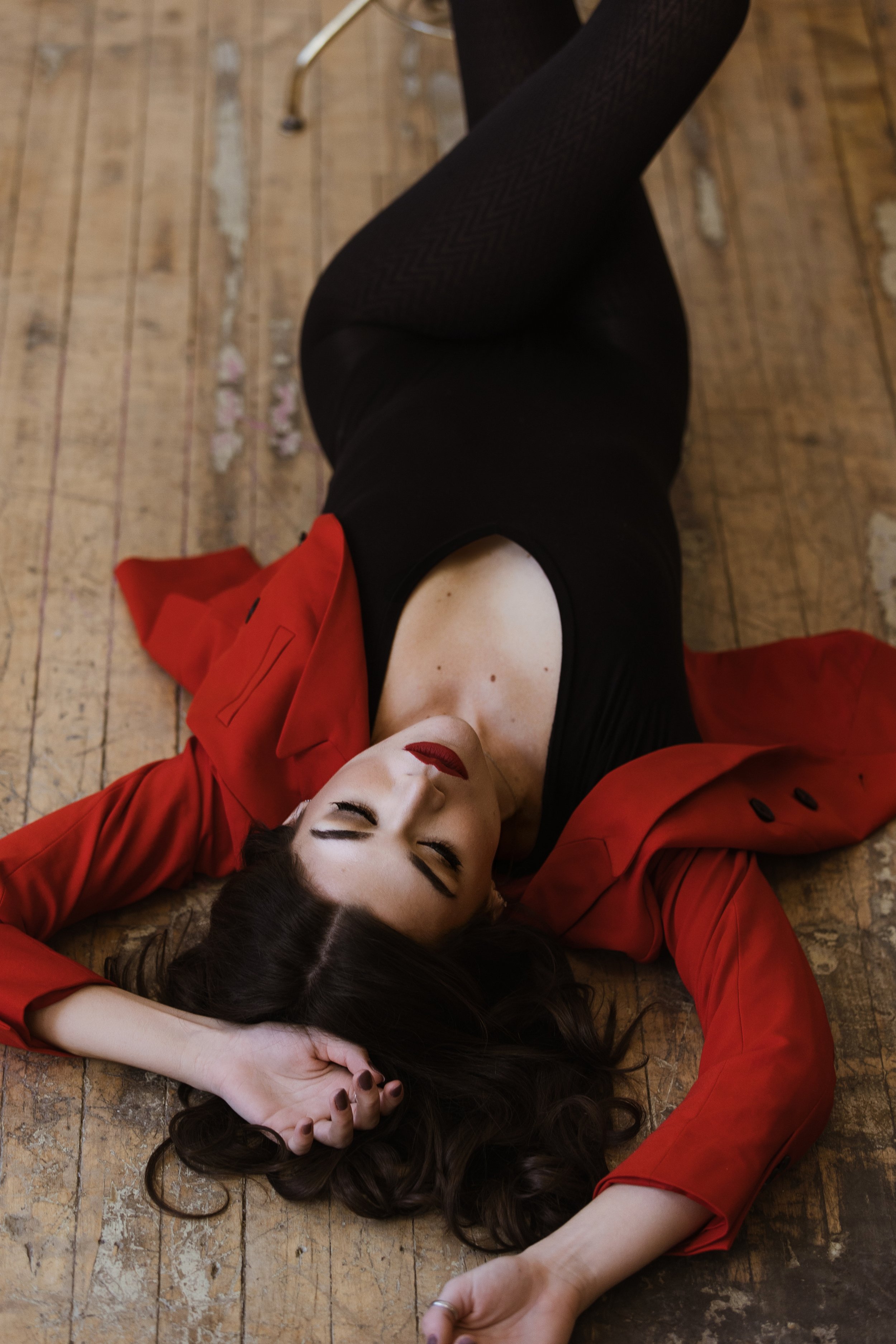A woman lying on a wooden floor with her eyes closed, wearing a black dress and a red jacket, with long dark hair spread out around her head.