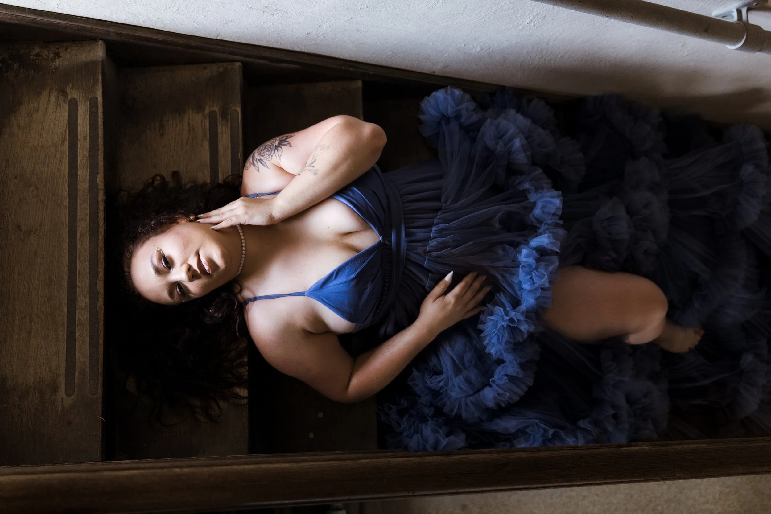 A woman lying on her side on a wooden floor near a staircase, wearing a blue dress with a ruffled skirt, accessorized with a pearl necklace, earrings, and a hairpin, with tattoos on her arm and posed with her hand resting on her face.