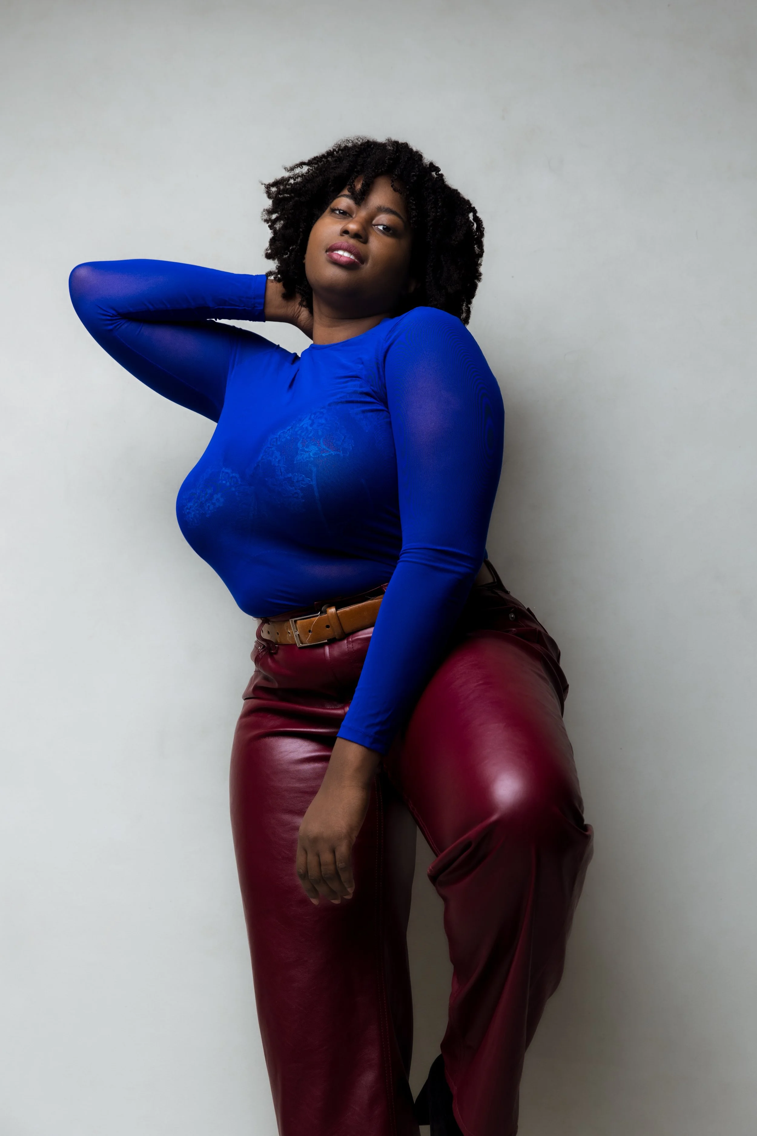 A woman with curly black hair wearing a royal blue long-sleeve top and maroon leather pants, standing against a plain light-colored wall.