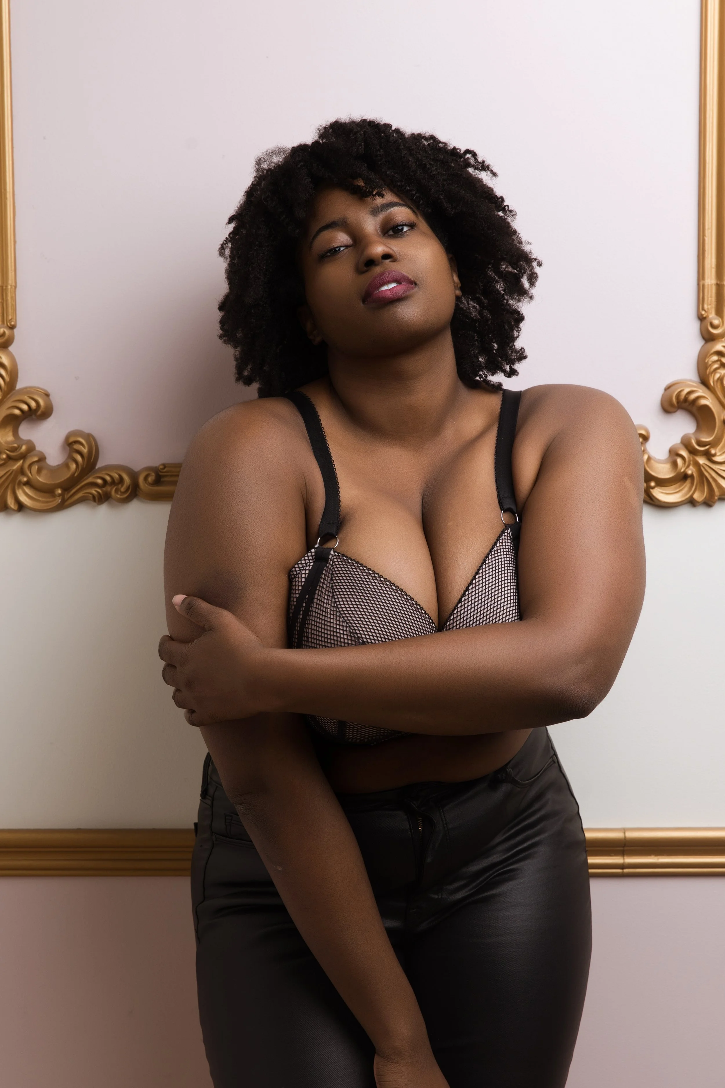 A confident woman with curly hair wearing a black netted top and black pants, standing against a wall with ornate gold decorations.