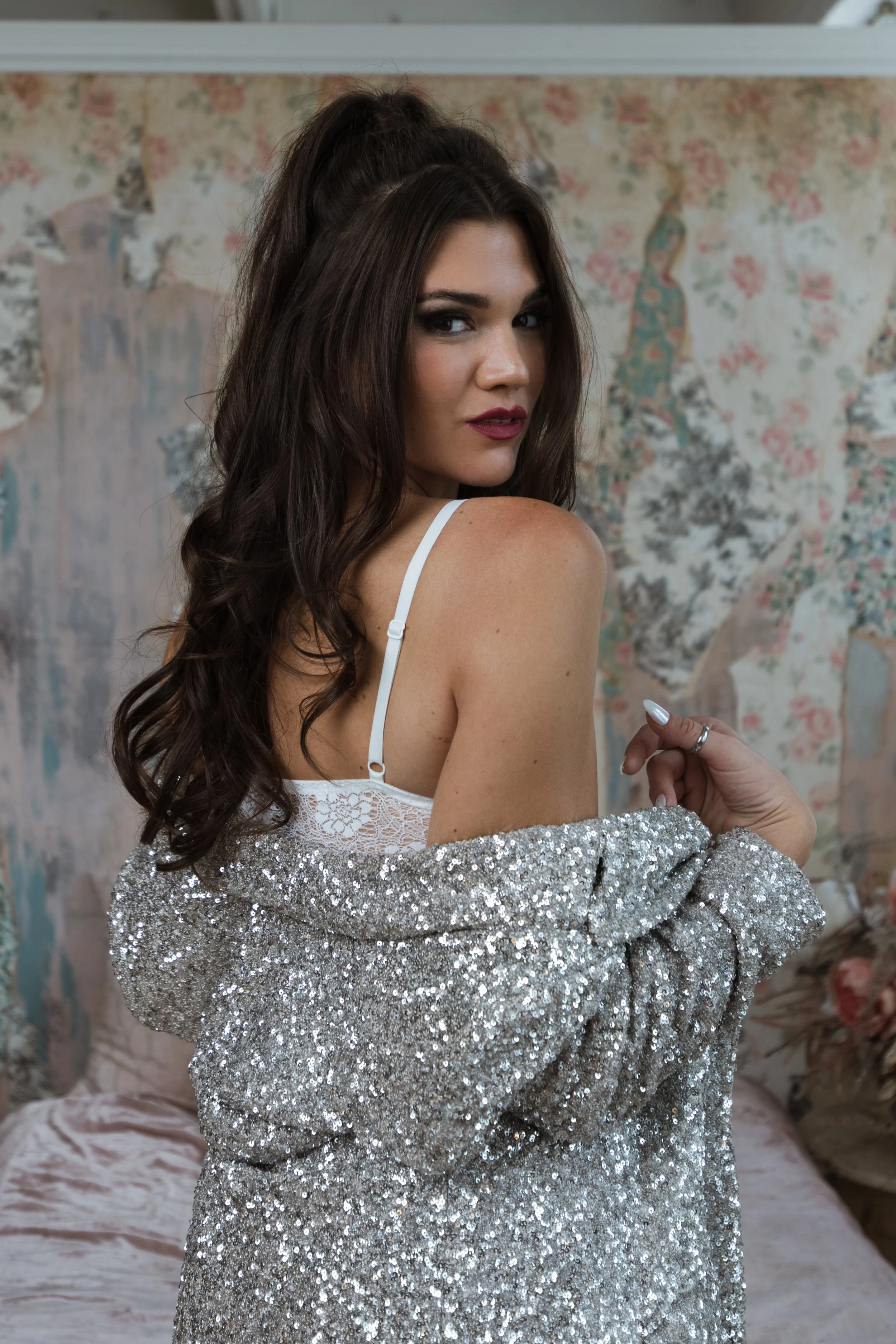 A woman with long, dark wavy hair, wearing a white lace bra and a sparkly silver jacket.