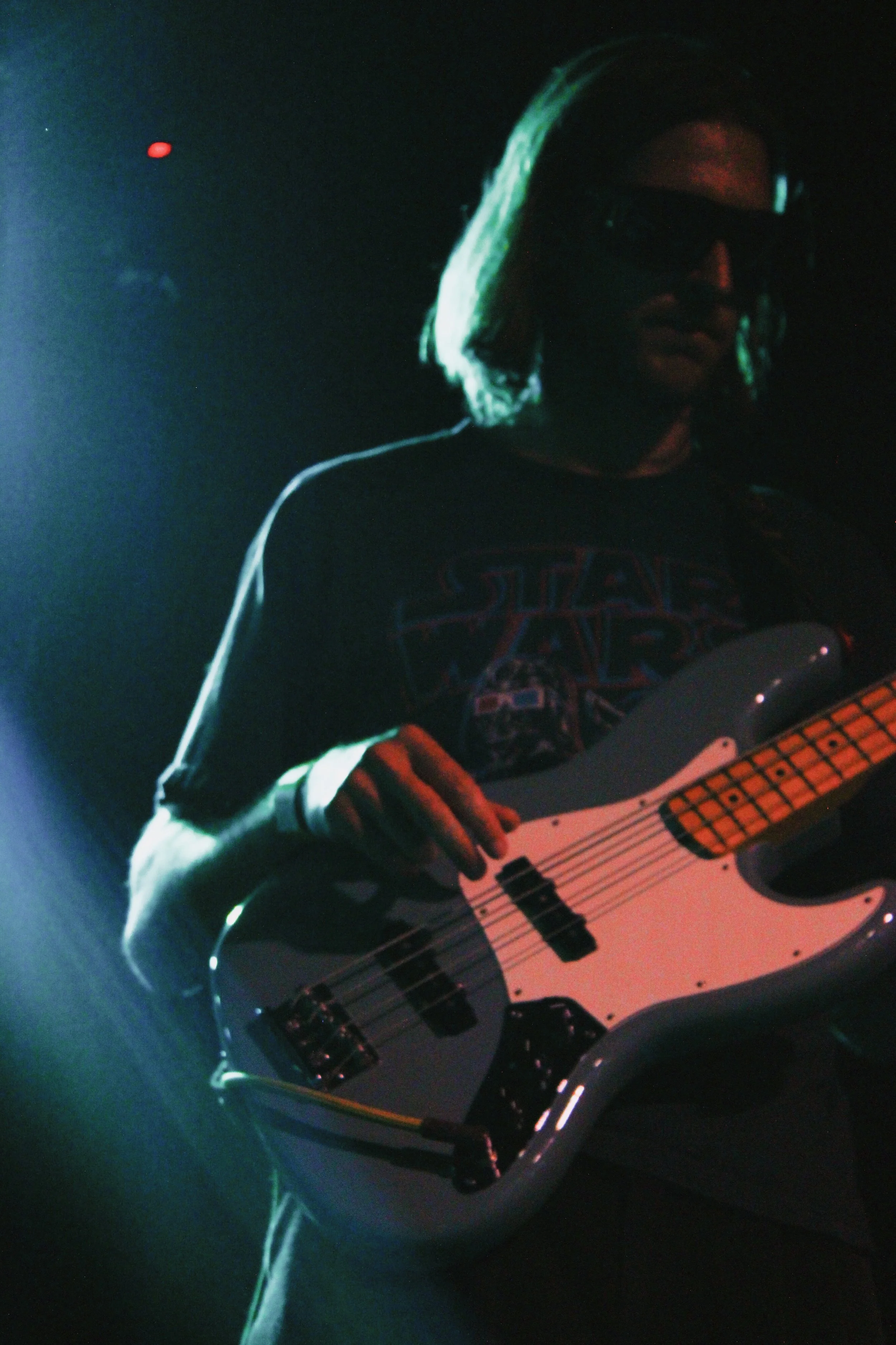 A person with long hair wearing sunglasses and a Star Wars T-shirt playing an electric bass guitar on stage with colorful lighting.