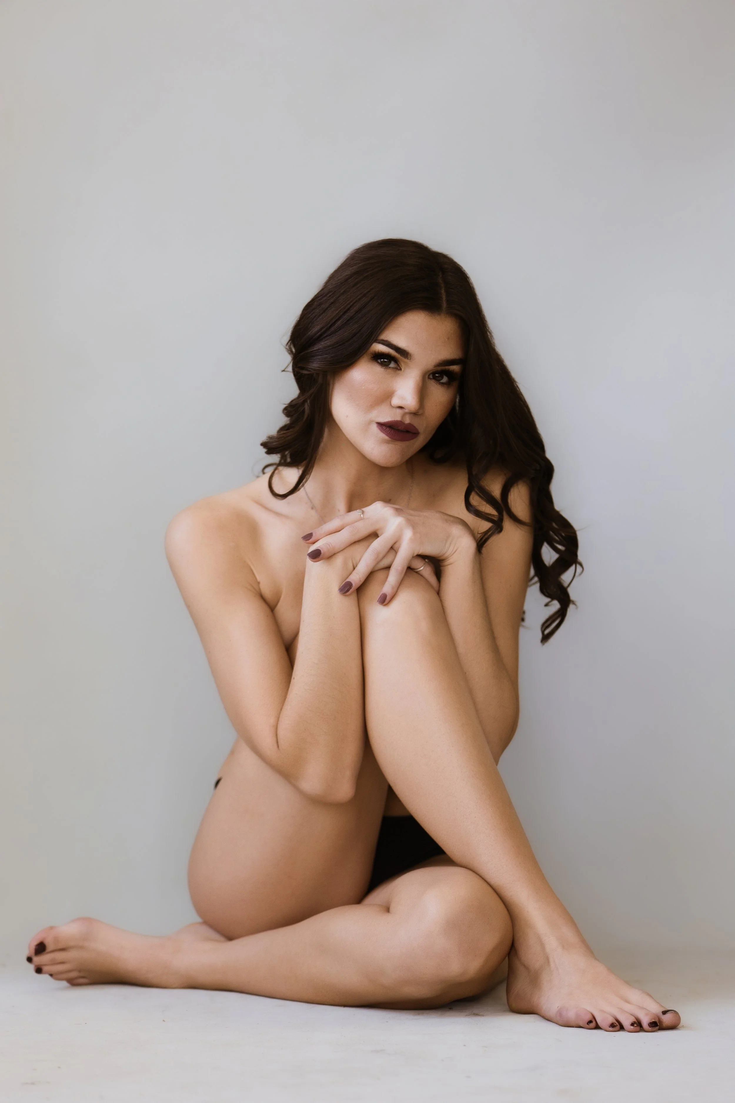 A woman sitting cross-legged against a plain light-colored wall, with long dark hair and dark lipstick, looking at the camera.