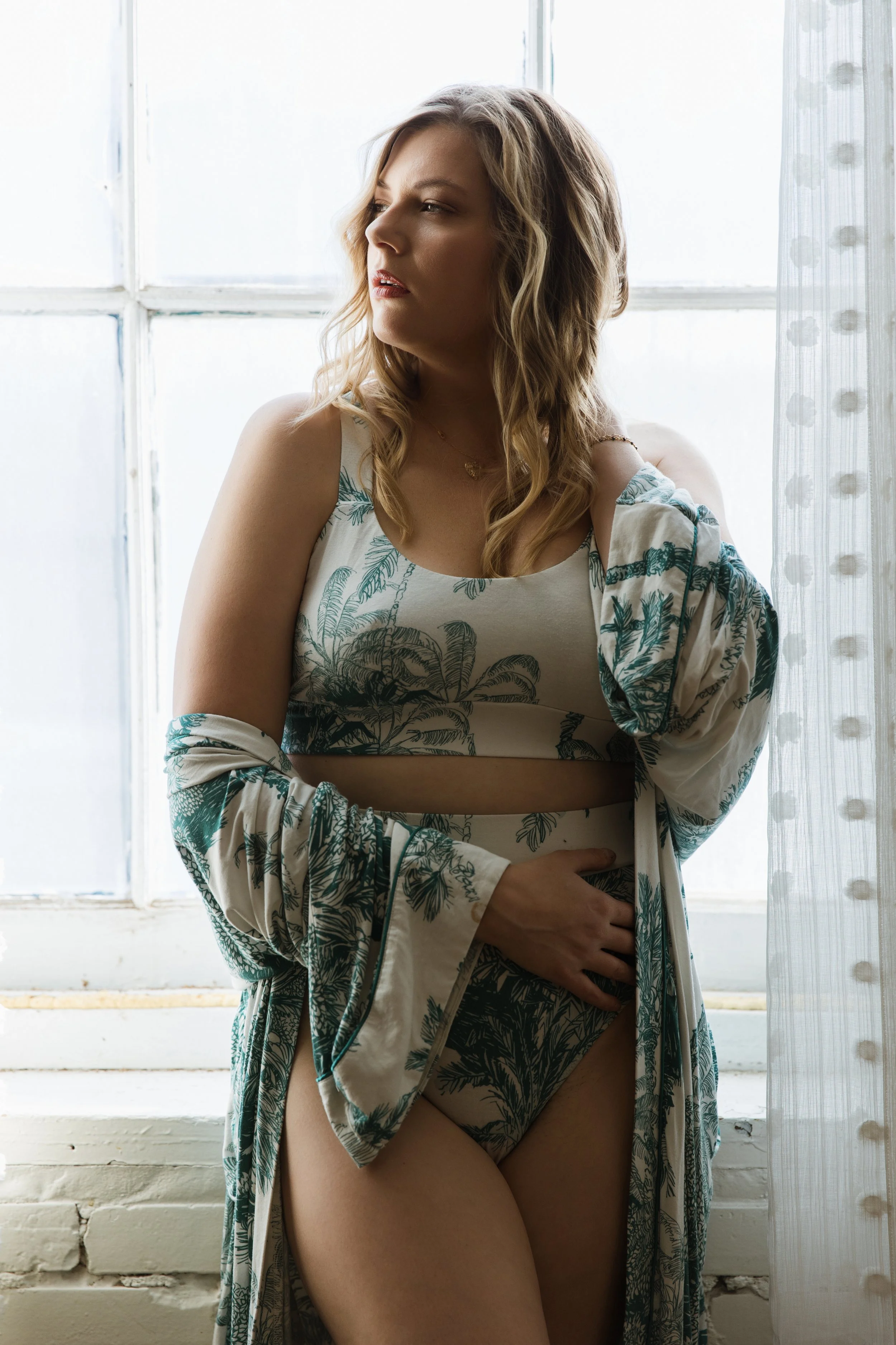 A woman standing by a window in natural light, wearing a botanical print lingerie set and a matching robe.