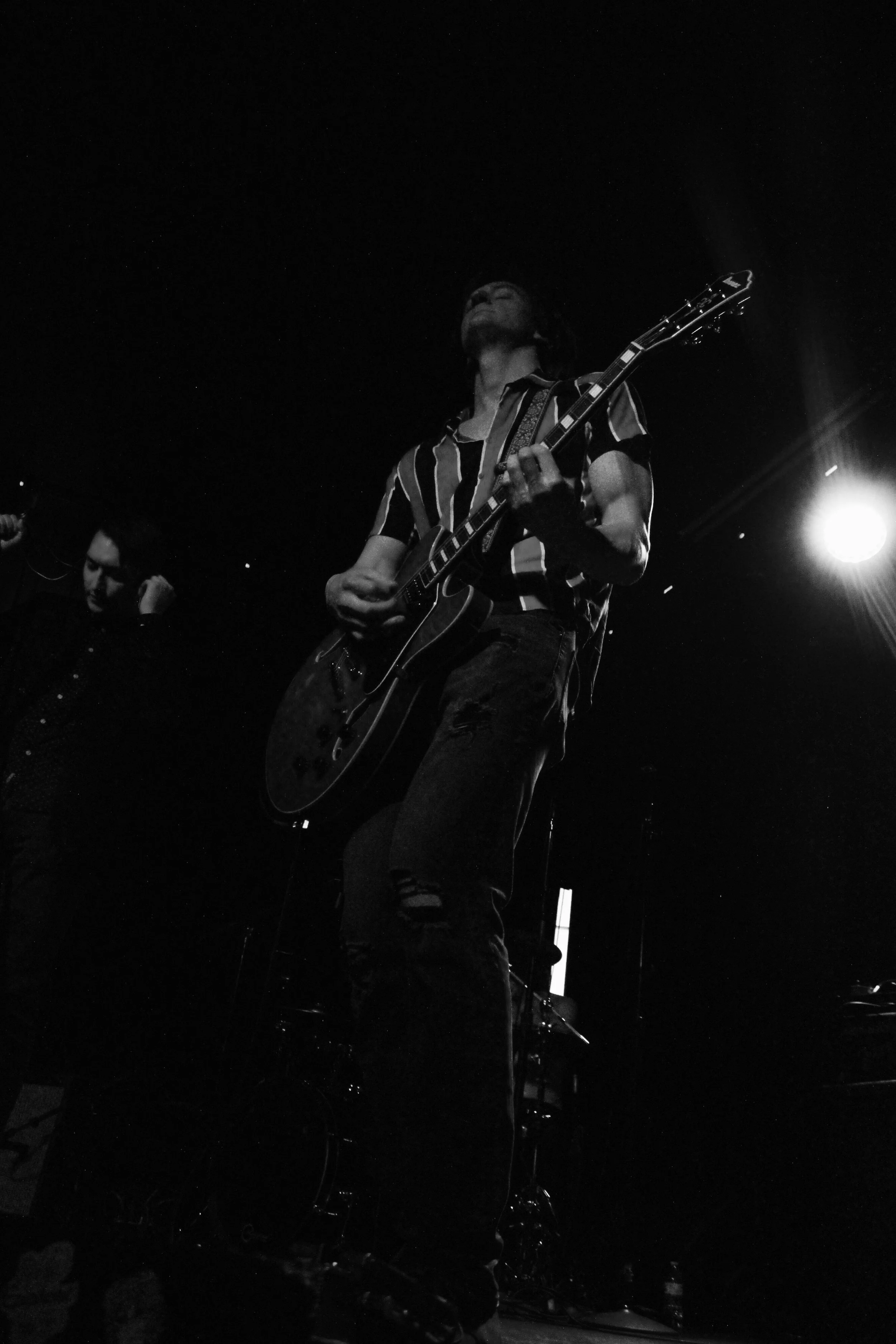 A black and white photo of a guitarist on stage playing an electric guitar, with a spotlight shining from the right side in a dark setting.