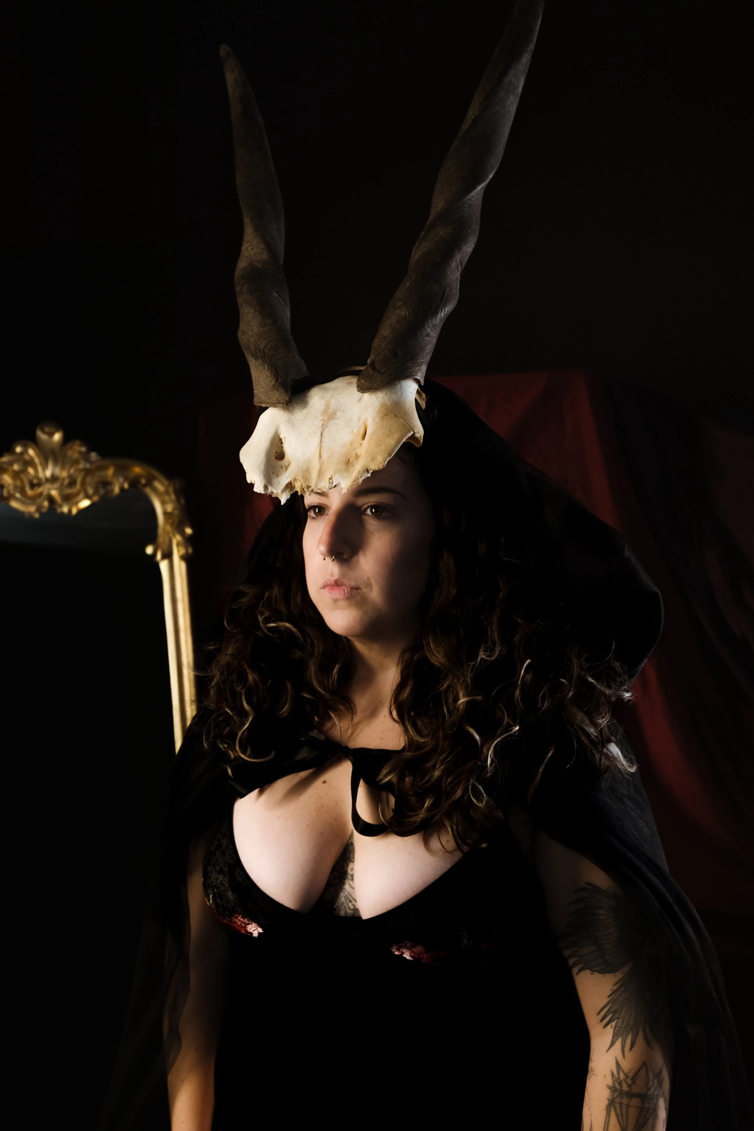 A woman with curly hair and tattoos wearing a black veil and cape, with a horned skull on her head, standing in front of a mirror with a gold frame, dark background, and red drapery.