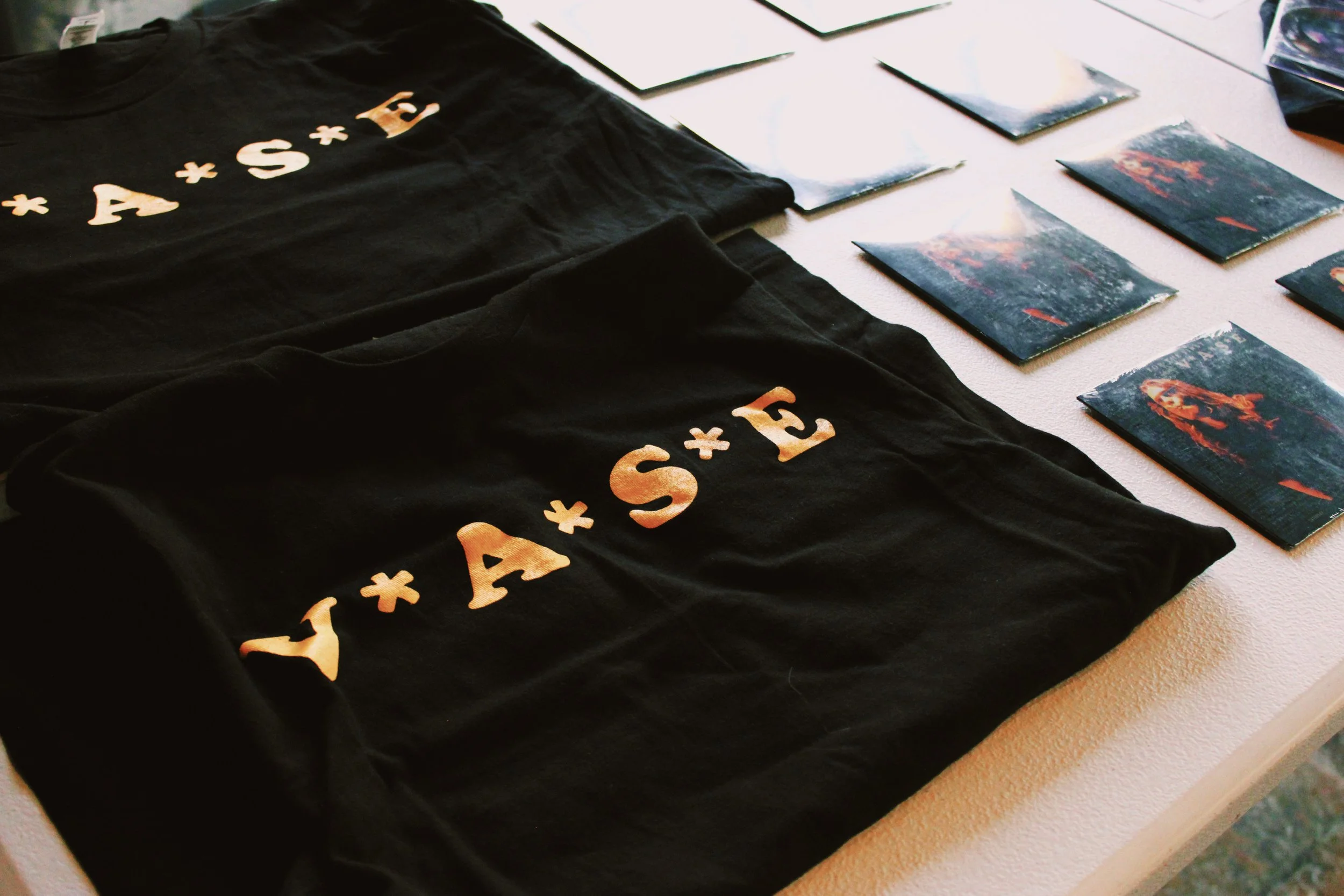 Black T-shirts with the text "VIVA + A + S + F" printed in gradient colors, lying on a table alongside several small square cards featuring artwork.