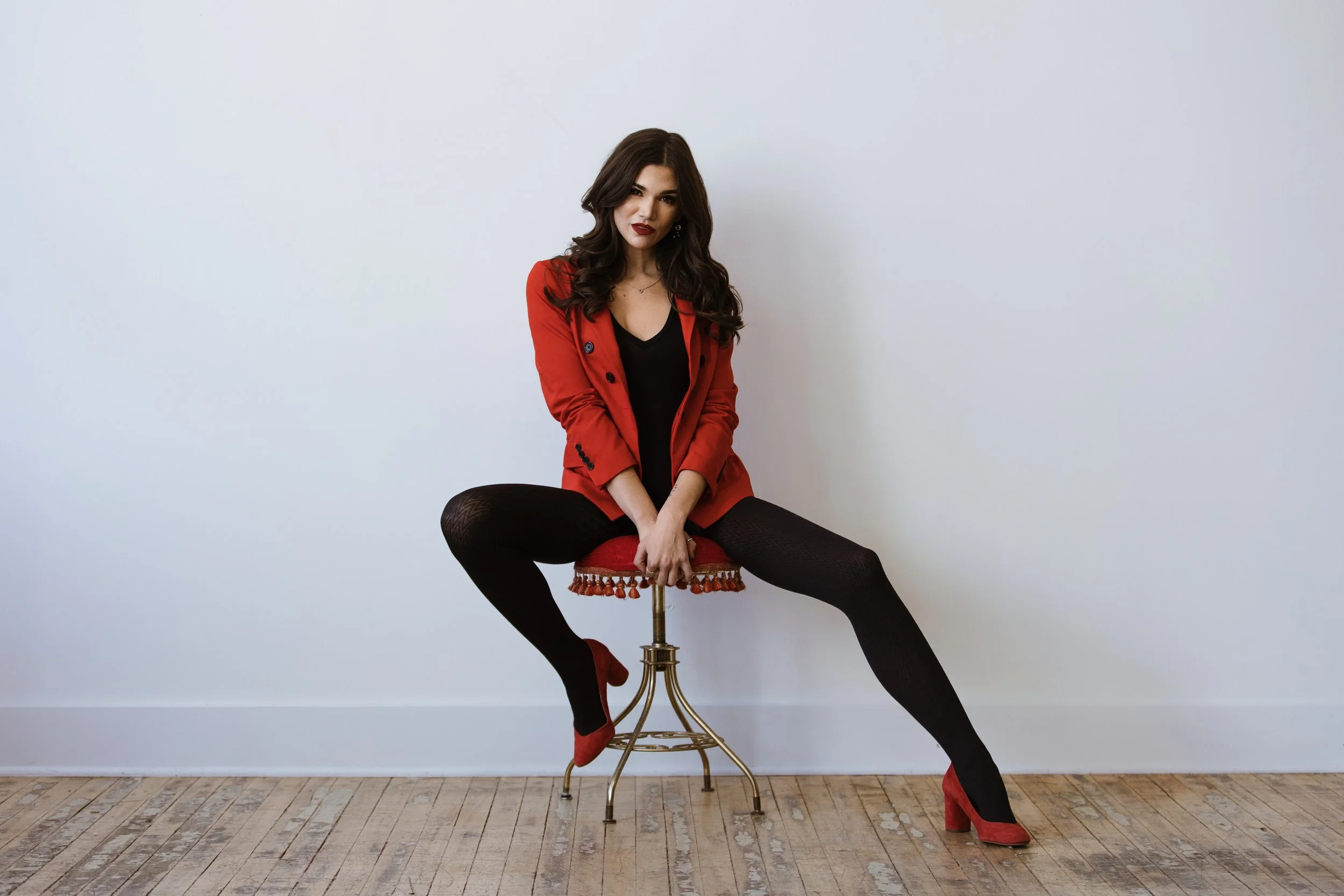 A woman sitting on a vintage stool with a red velvet cushion and fringe trim, against a plain white wall. She is wearing a red blazer, black top, black tights, and red high heels, and has long dark hair and red lipstick.
