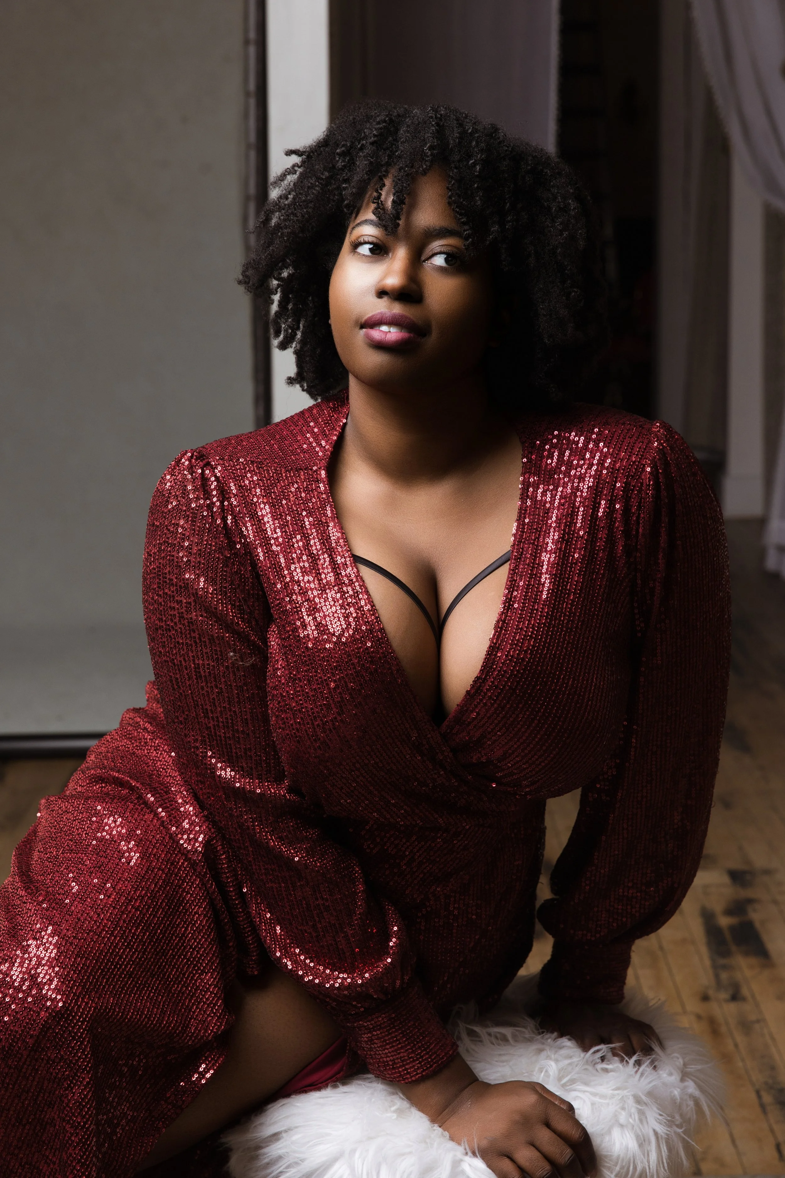 A woman with natural curly hair poses indoors wearing a shimmering red dress with a plunging neckline and black strappy details, against a neutral background with wooden flooring.