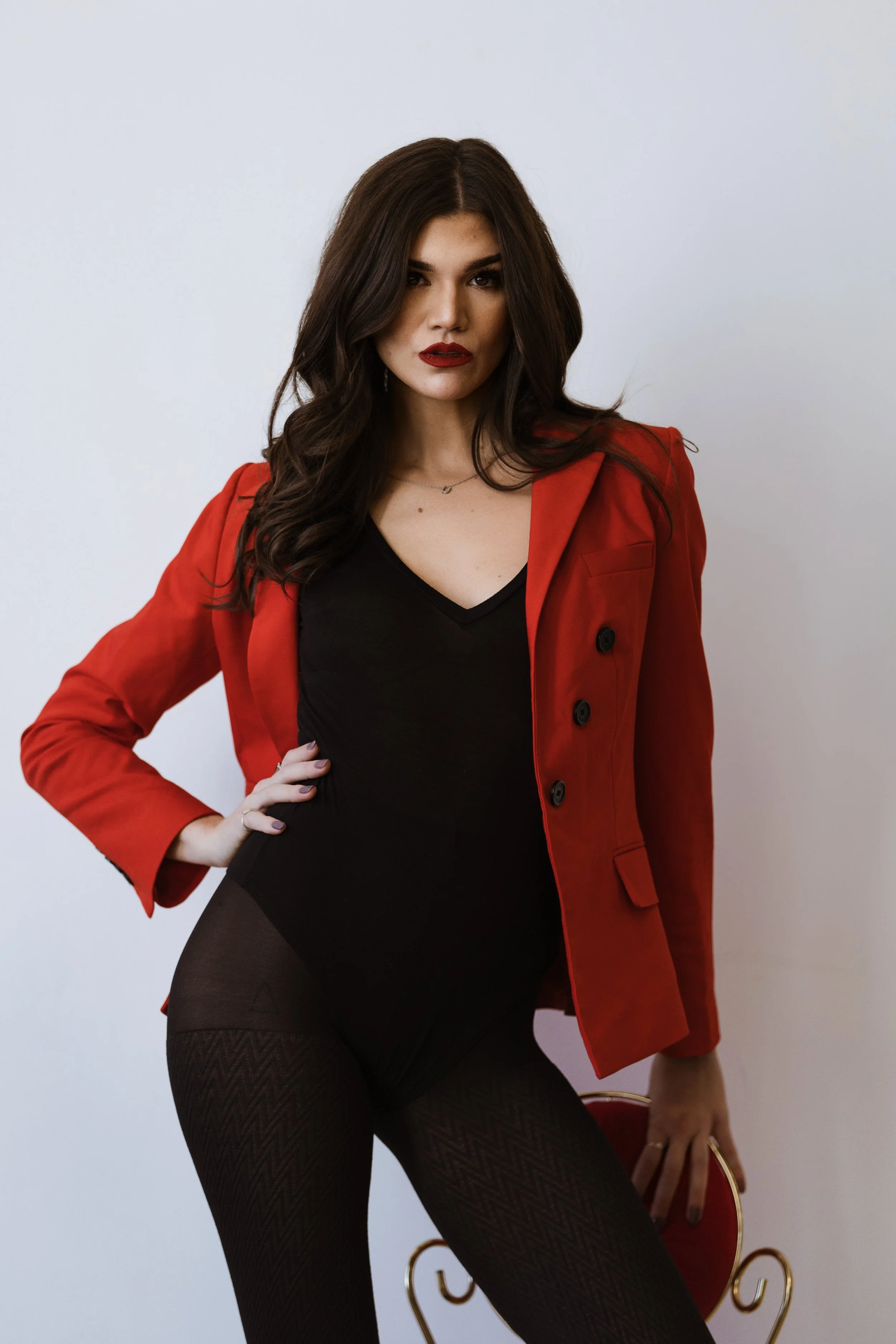 A woman with long dark hair wearing a black top, red blazer, and black patterned tights, standing against a plain light-colored wall.