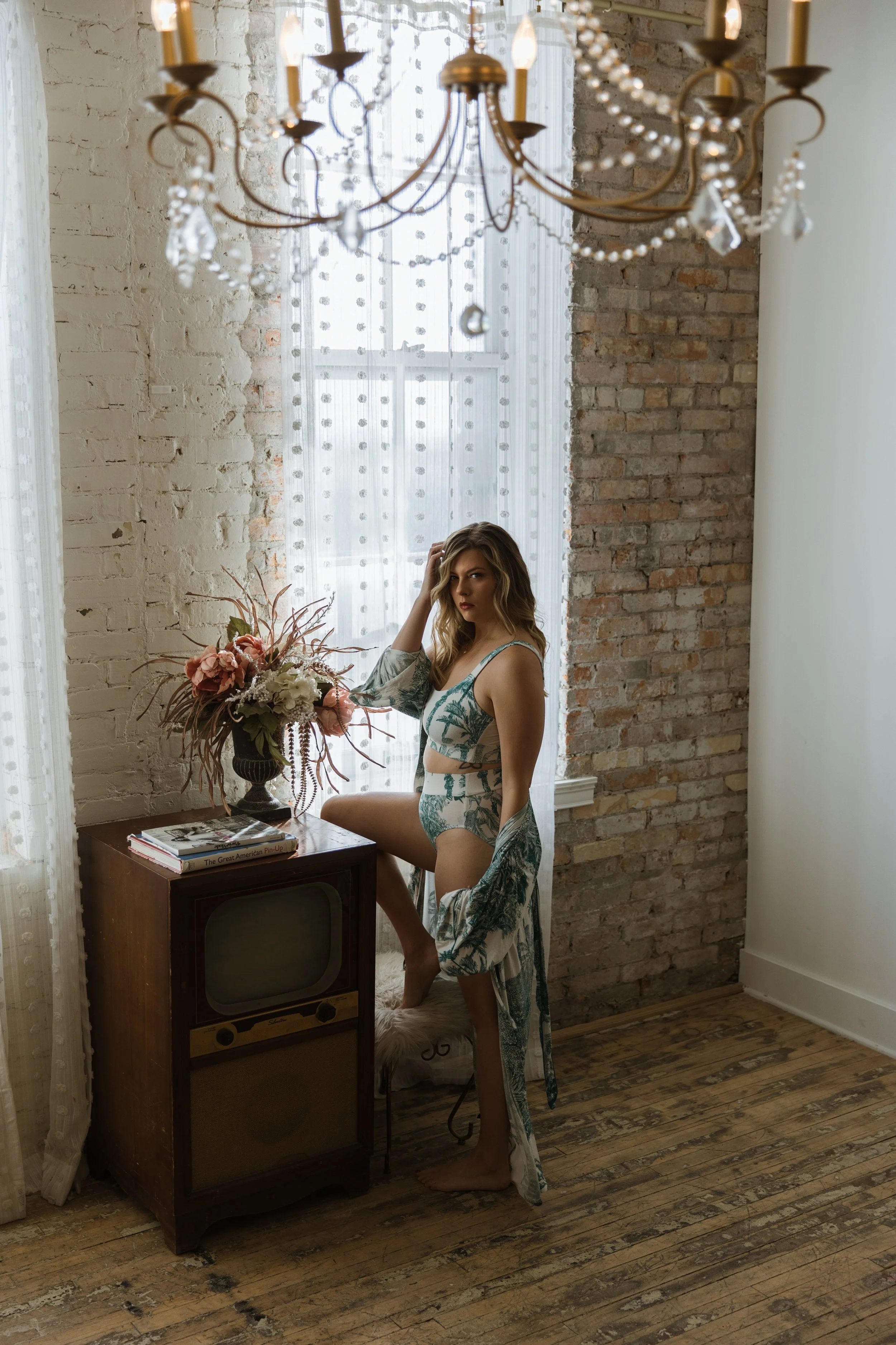 A woman in vintage-style lingerie and a matching robe stands in a rustic room with exposed brick walls. She is posed next to a small table with a vintage television and a large floral arrangement. The room has sheer curtains, a brick wall backdrop, a