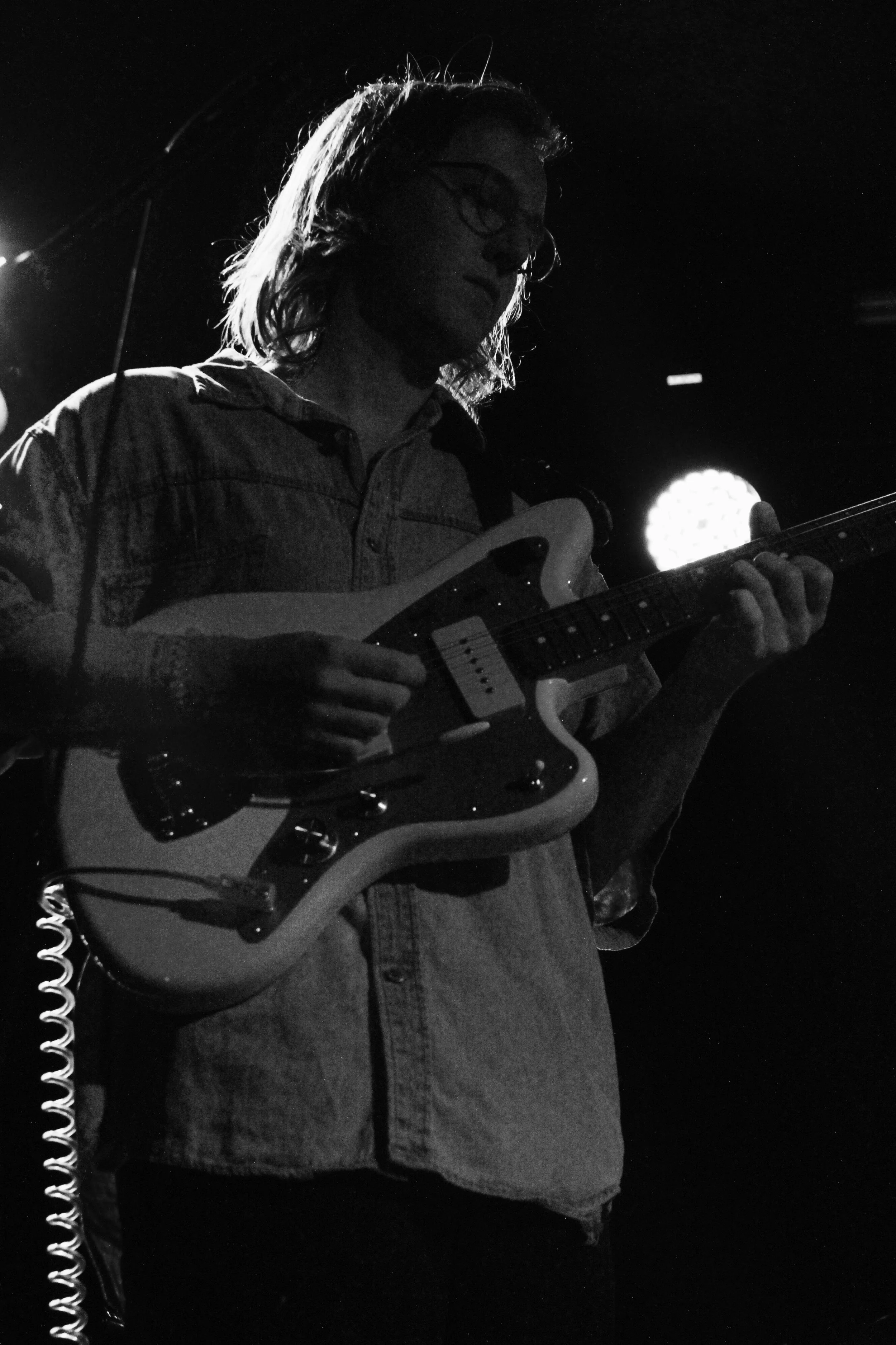 A person playing an electric guitar on stage in a black and white photo, with a bright light behind them.