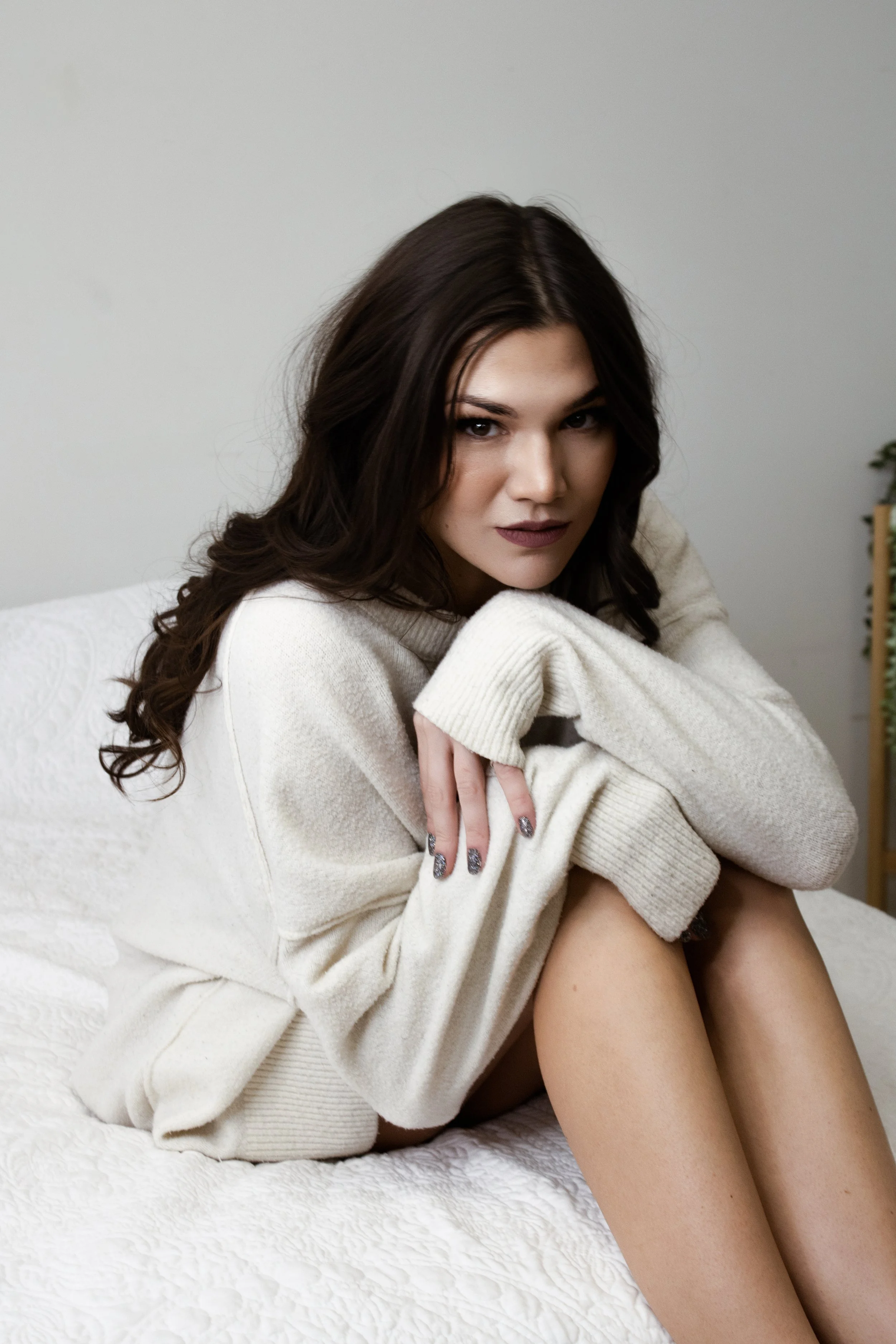 A woman with brown hair and dark lipstick sitting on a bed in a cream sweater, looking at the camera.
