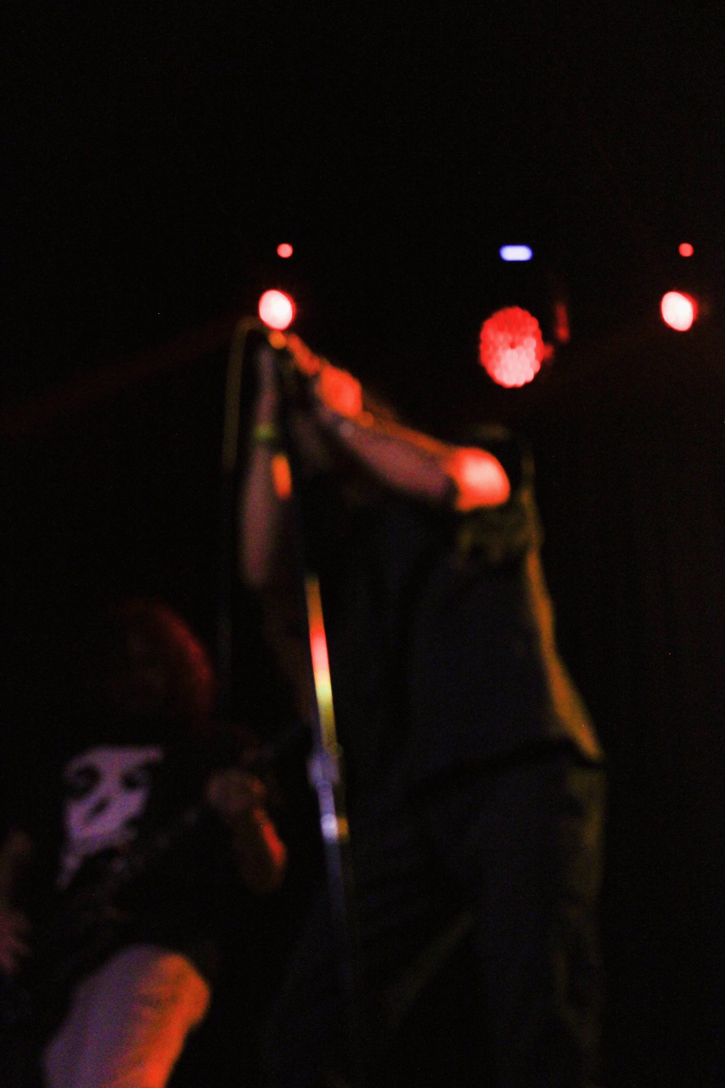 A person, partially visible, wearing a mask, holding a microphone stand, with stage lights in the background.
