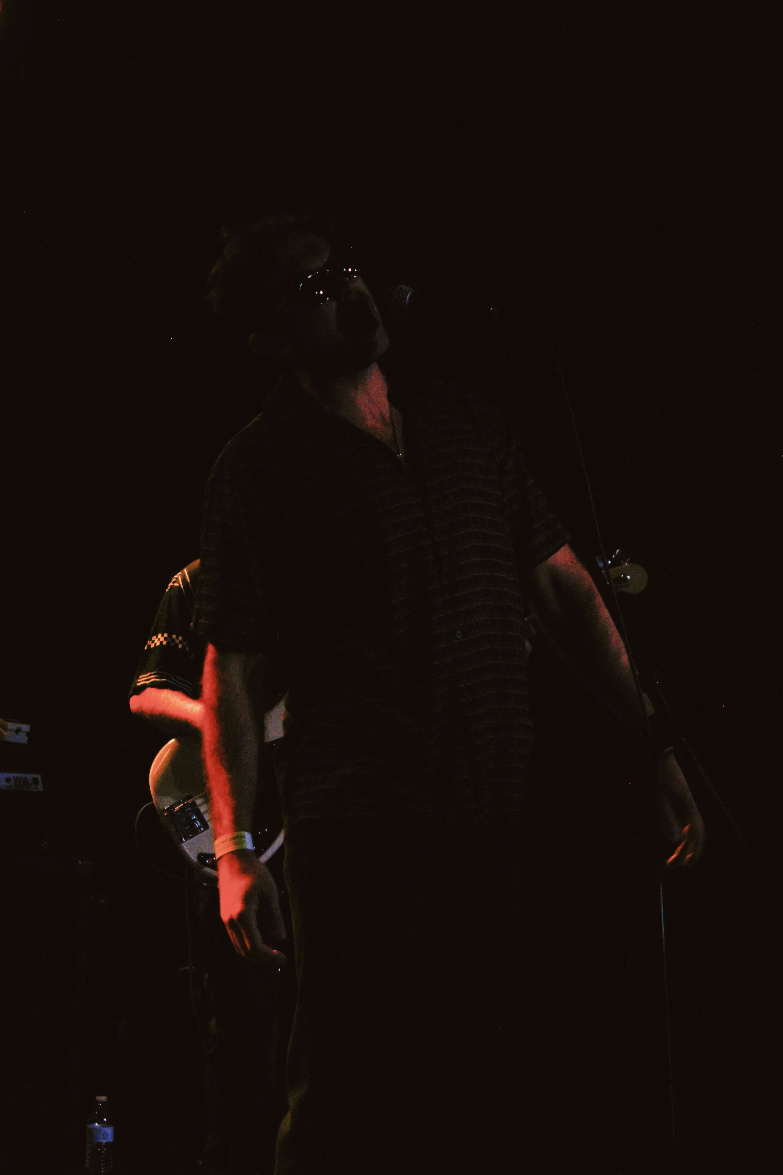 A man standing on a dark stage with a microphone, illuminated by dim, colored stage lights.