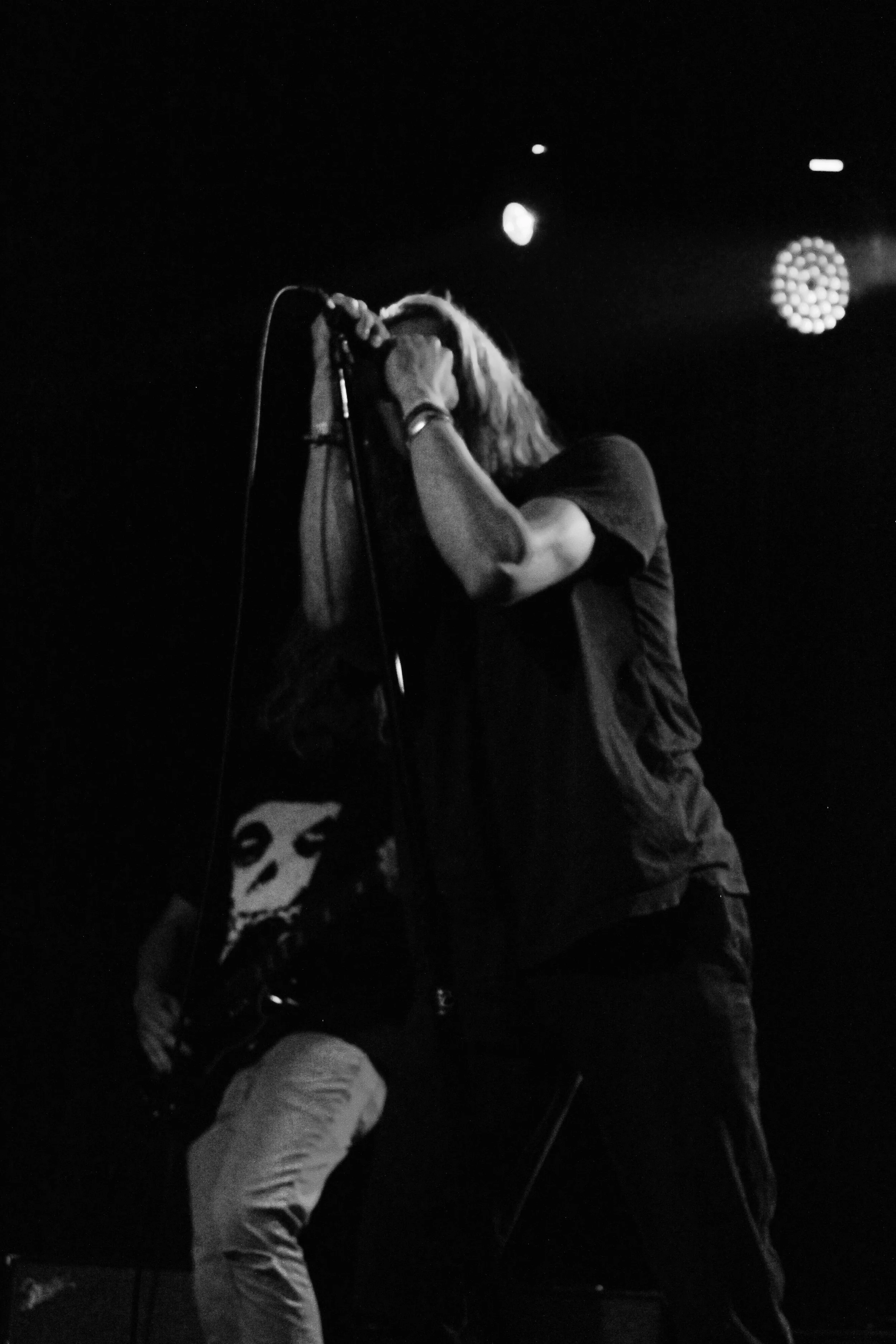 A black and white photo of a band performing on stage, with a vocalist holding a microphone and a guitarist wearing a skull t-shirt in the background.