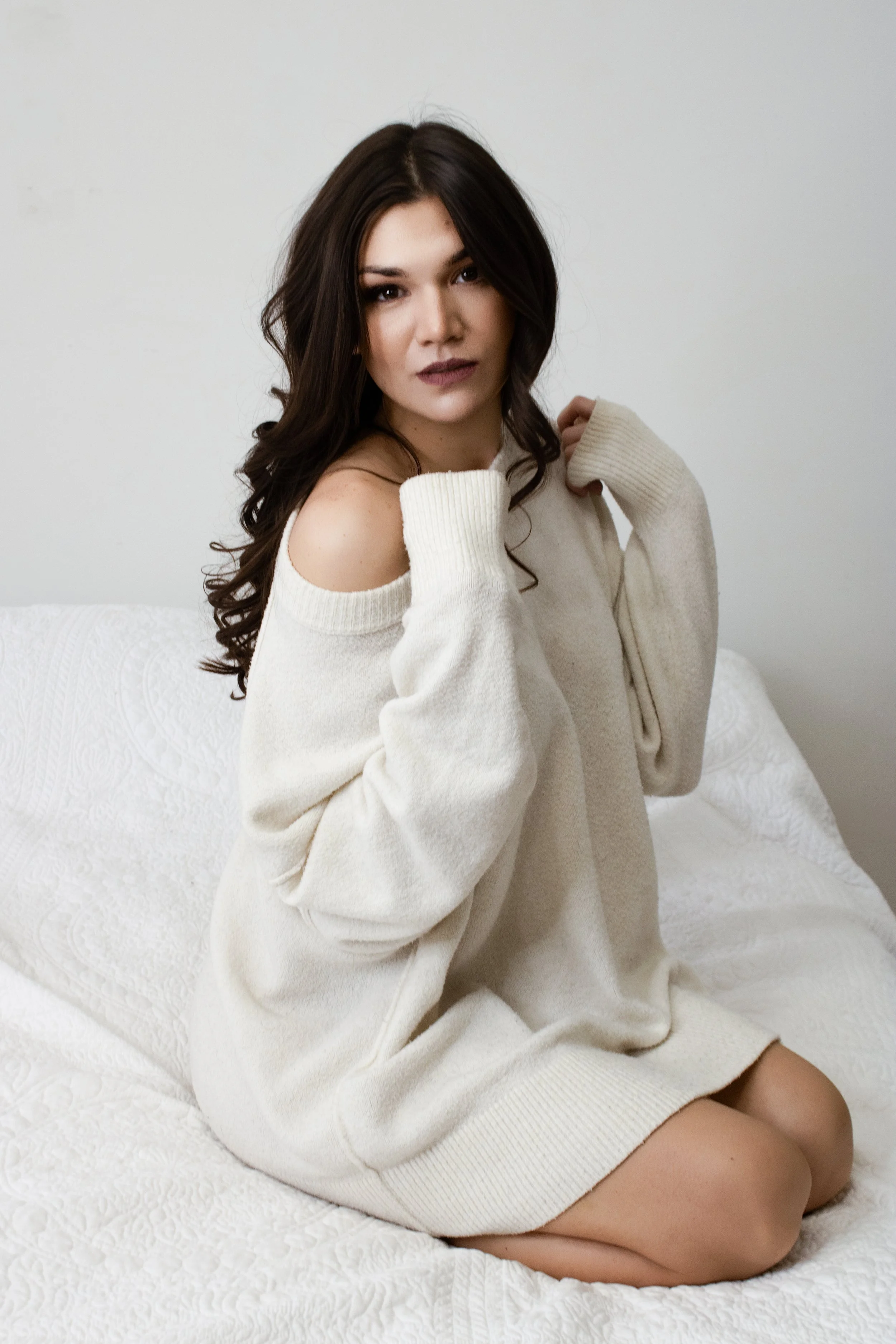 A young woman with dark wavy hair sitting on a bed in a cream sweater that slips off her shoulders, looking at the camera.