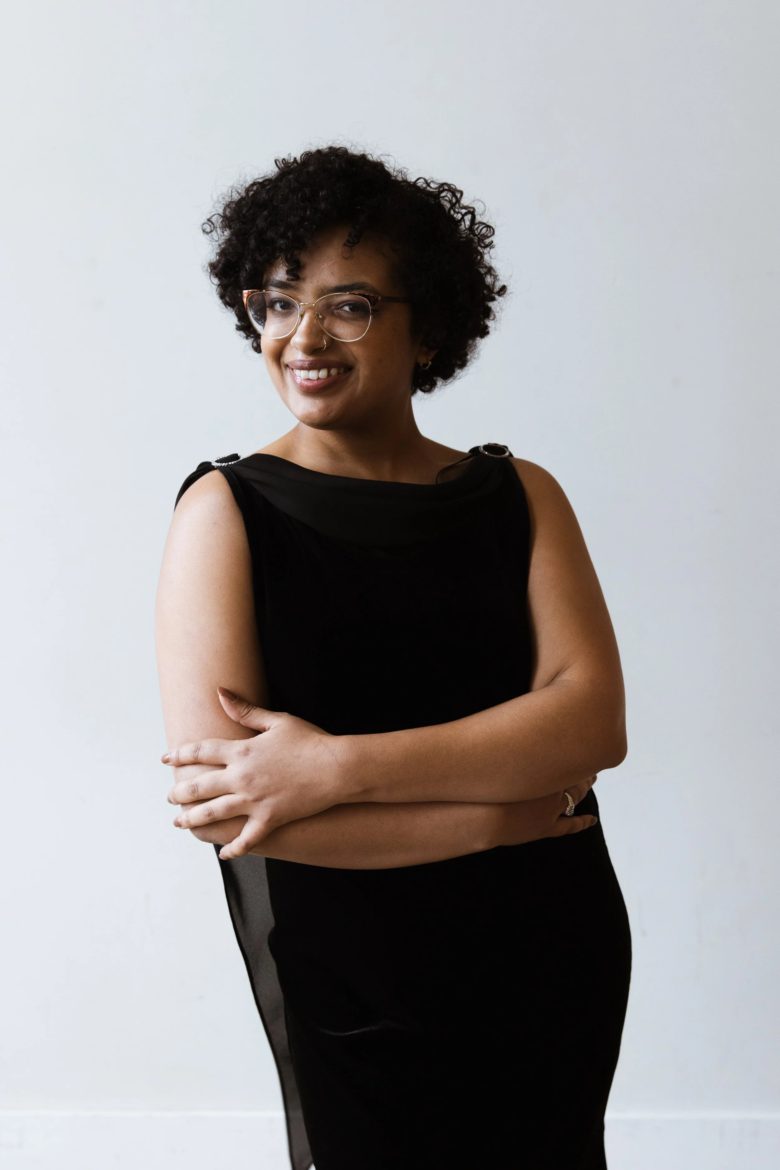 Portrait of a smiling woman with curly hair, glasses, wearing a sleeveless black dress, standing against a plain white background.