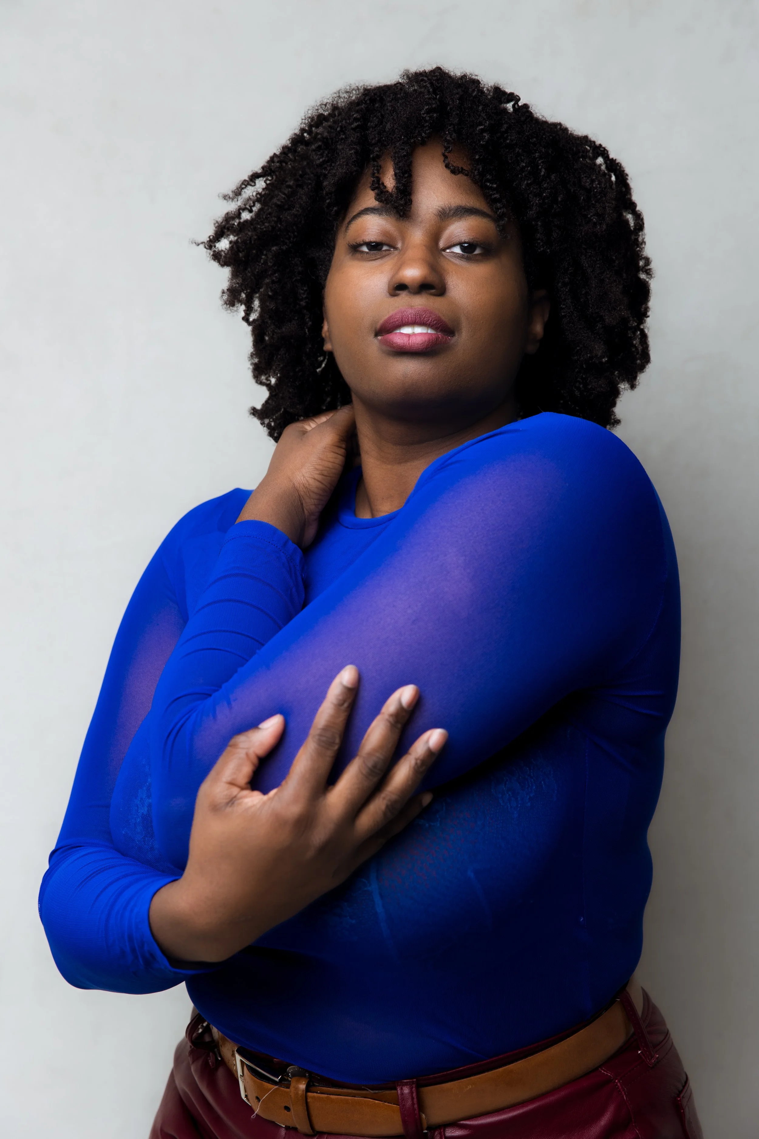 Portrait of a Black woman with curly hair wearing a blue long-sleeve top and maroon pants, standing against a plain background.