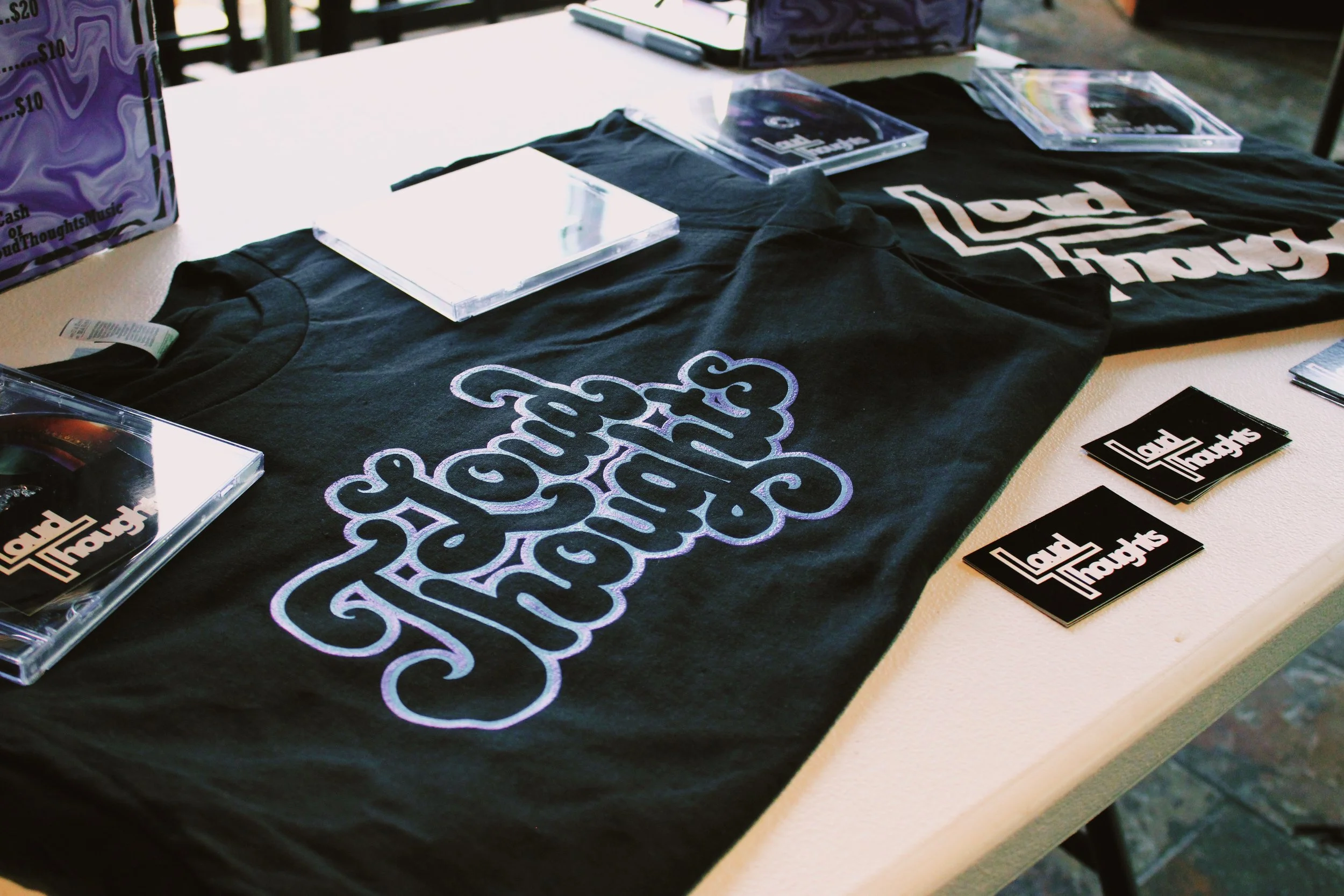Table with black T-shirts that have 'Land of Thought' embroidered on them, along with CDs in jewel cases and black cards displaying the logo.